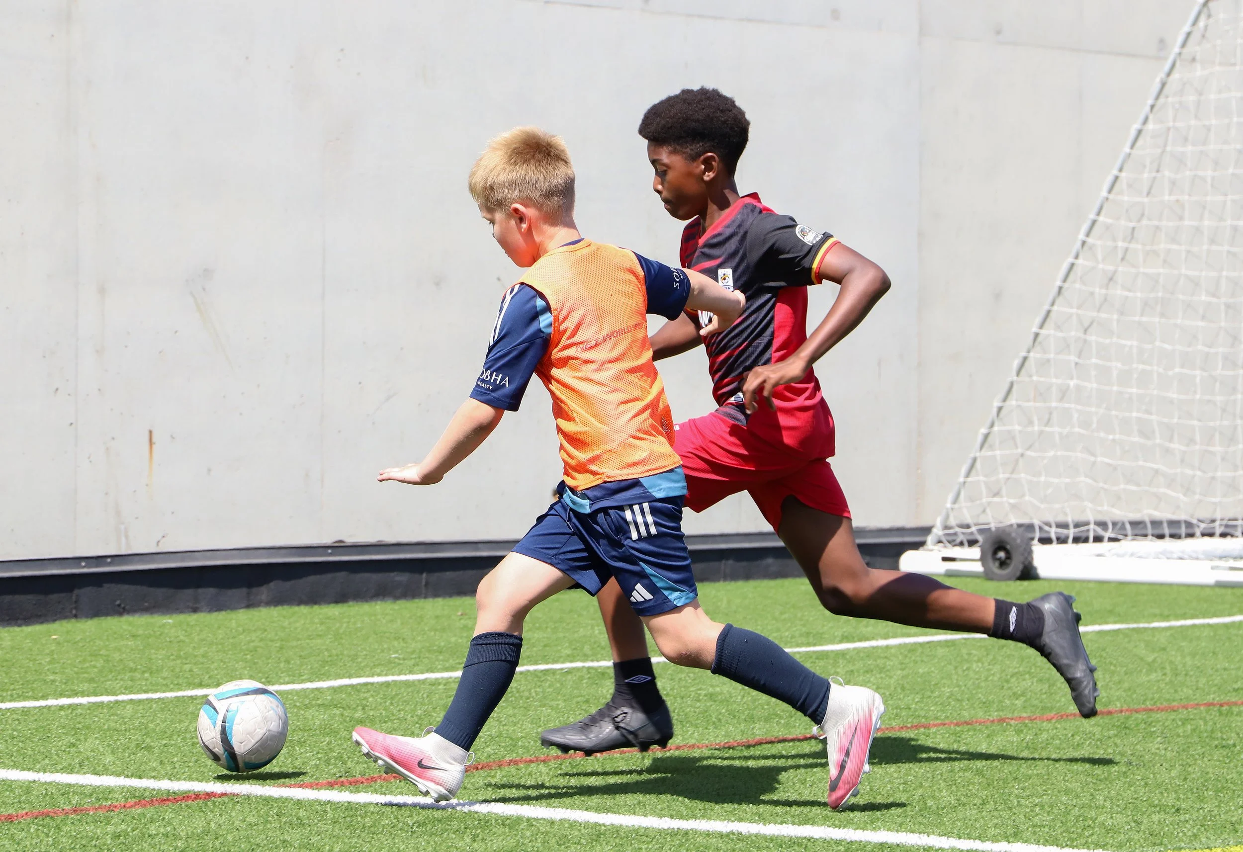 Two children playing football on a green field, one with blond hair in an orange and blue jersey, the other with black hair in a black and red jersey, chasing after a footballnear a goal.