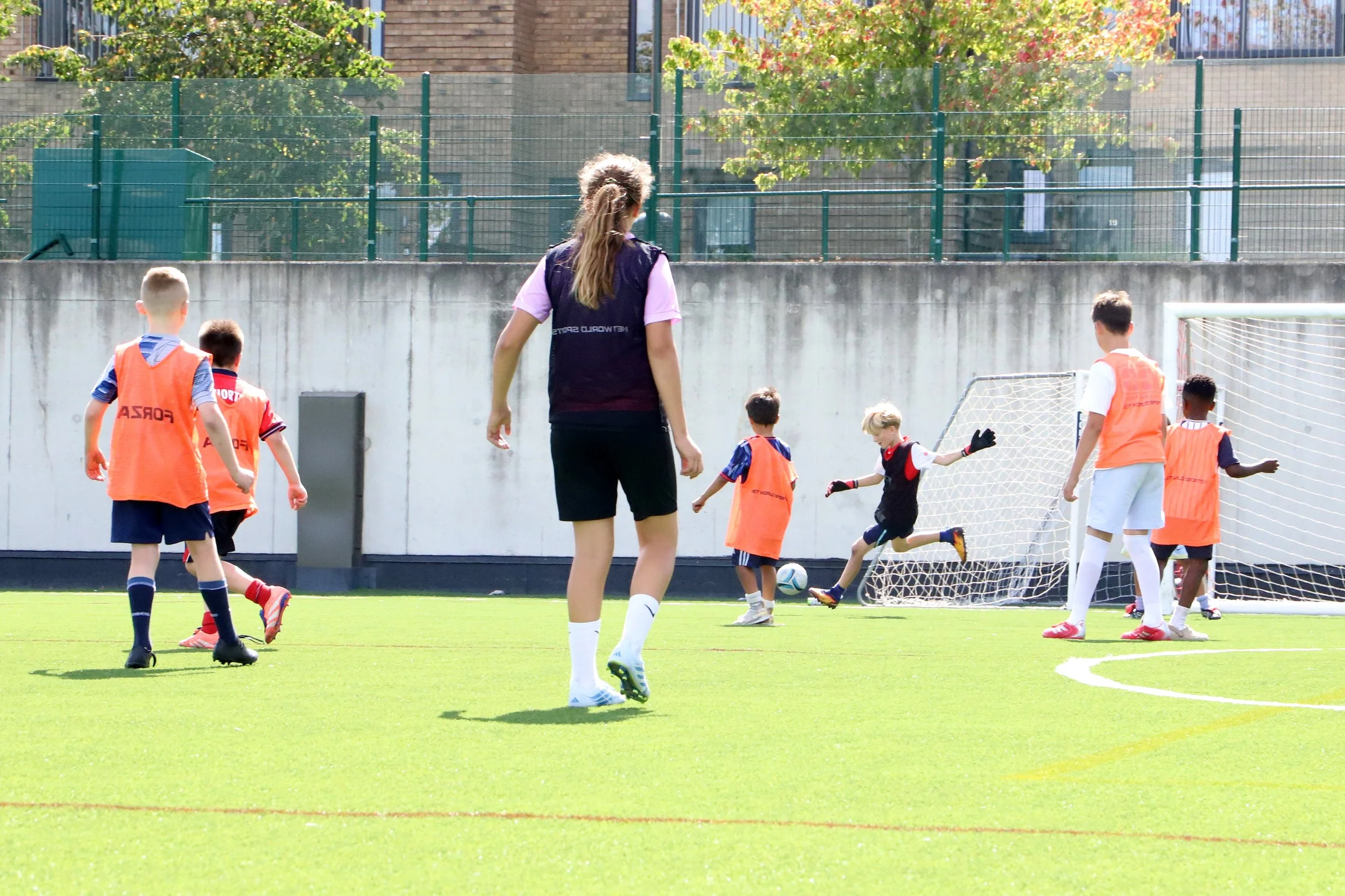 Children playing football on a green field with a coach or instructor observing, under a sunny sky.