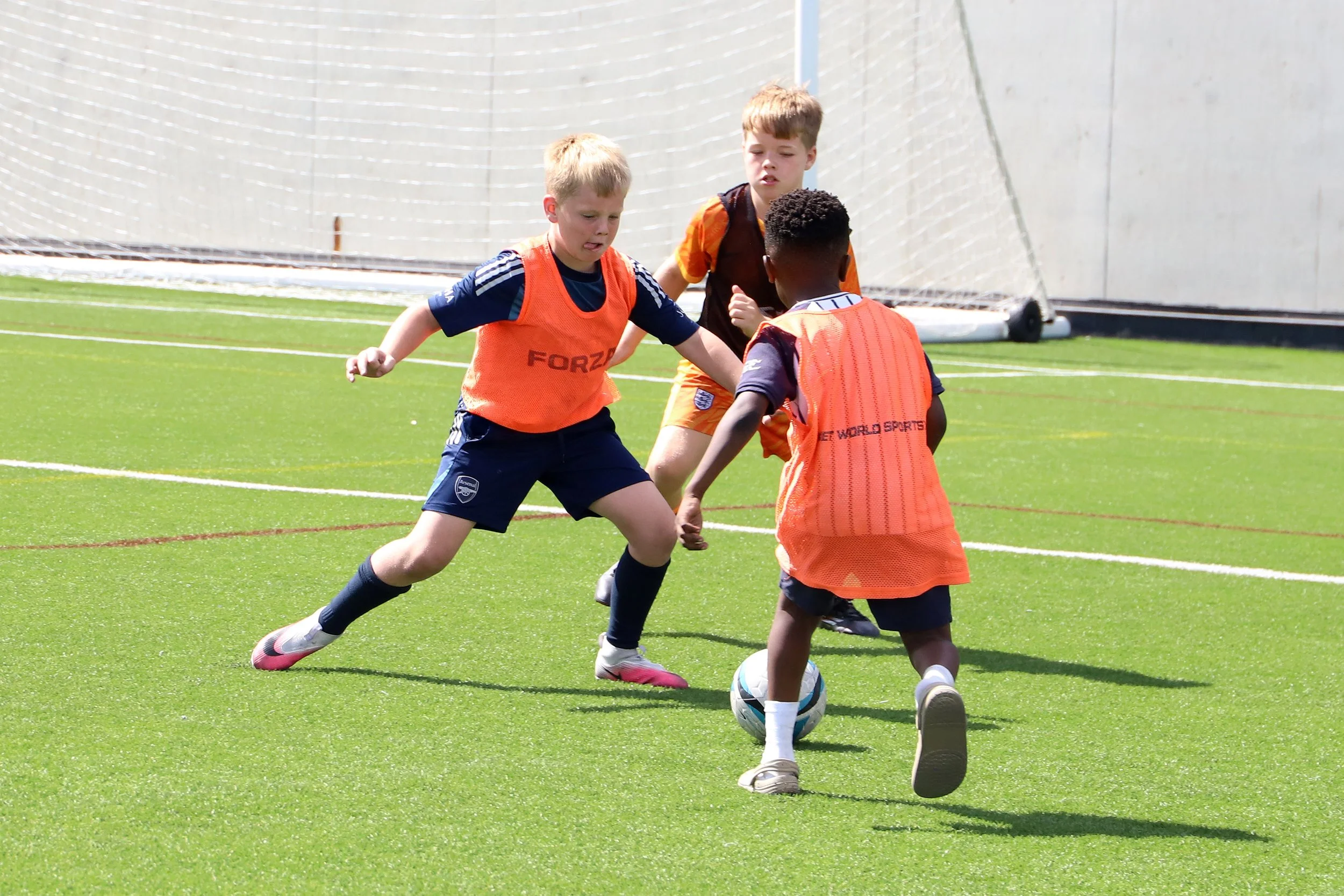 Three young boys playing football on a green field, two wearing orange vests and one wearing a dark shirt. All are focused on the ball, with a goal net in the background.