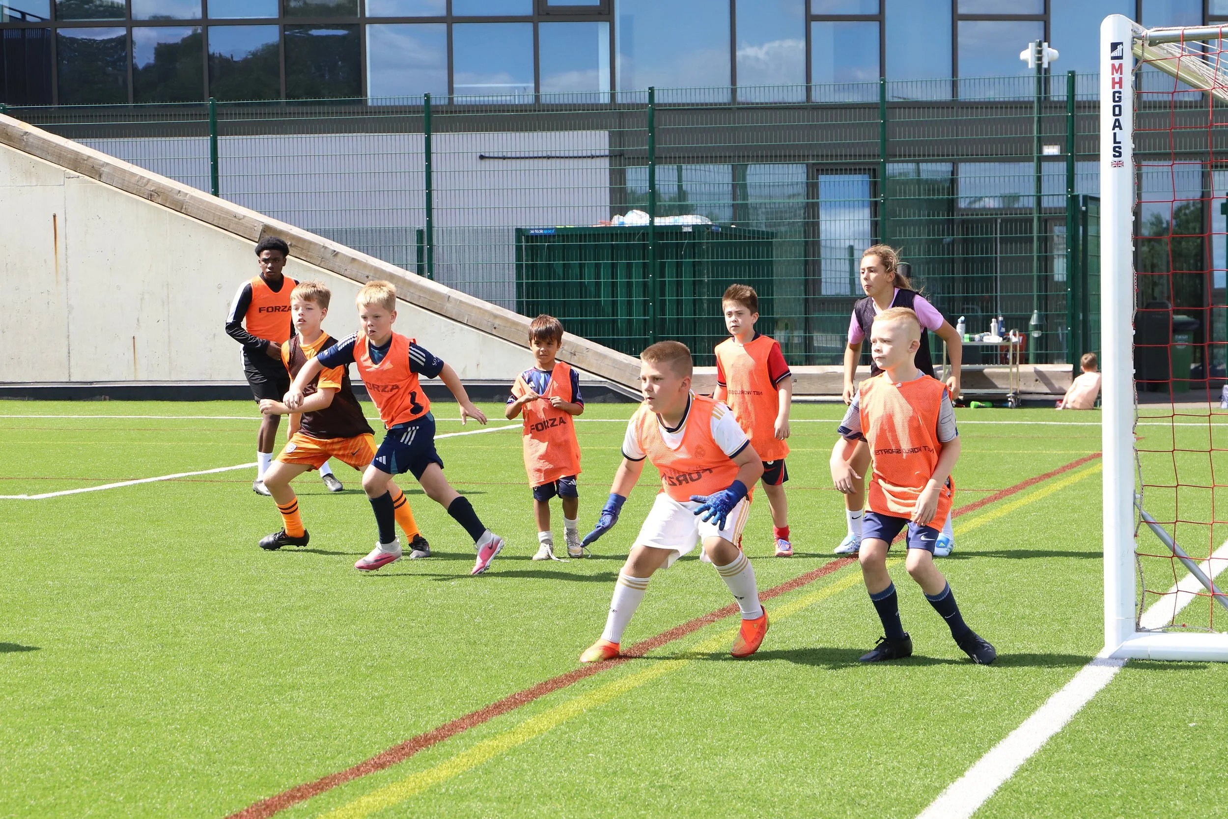Children playing football on a green field near the goal post with a glass building in the background.