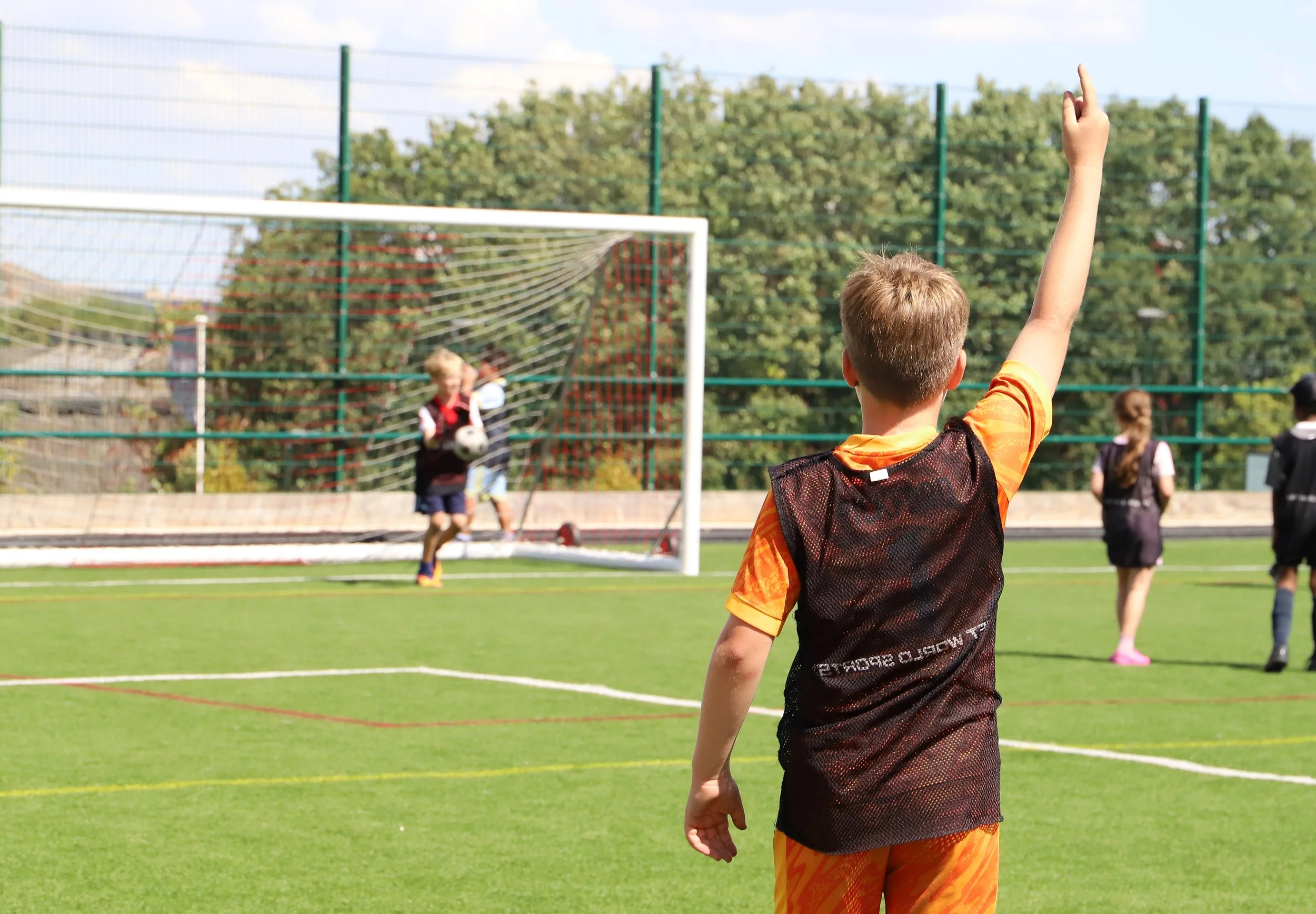 A young boy in an orange sports jersey with a black vest is seen raising his hand during a football game, facing a goal with children, including a girl with blonde hair, playing in the background.