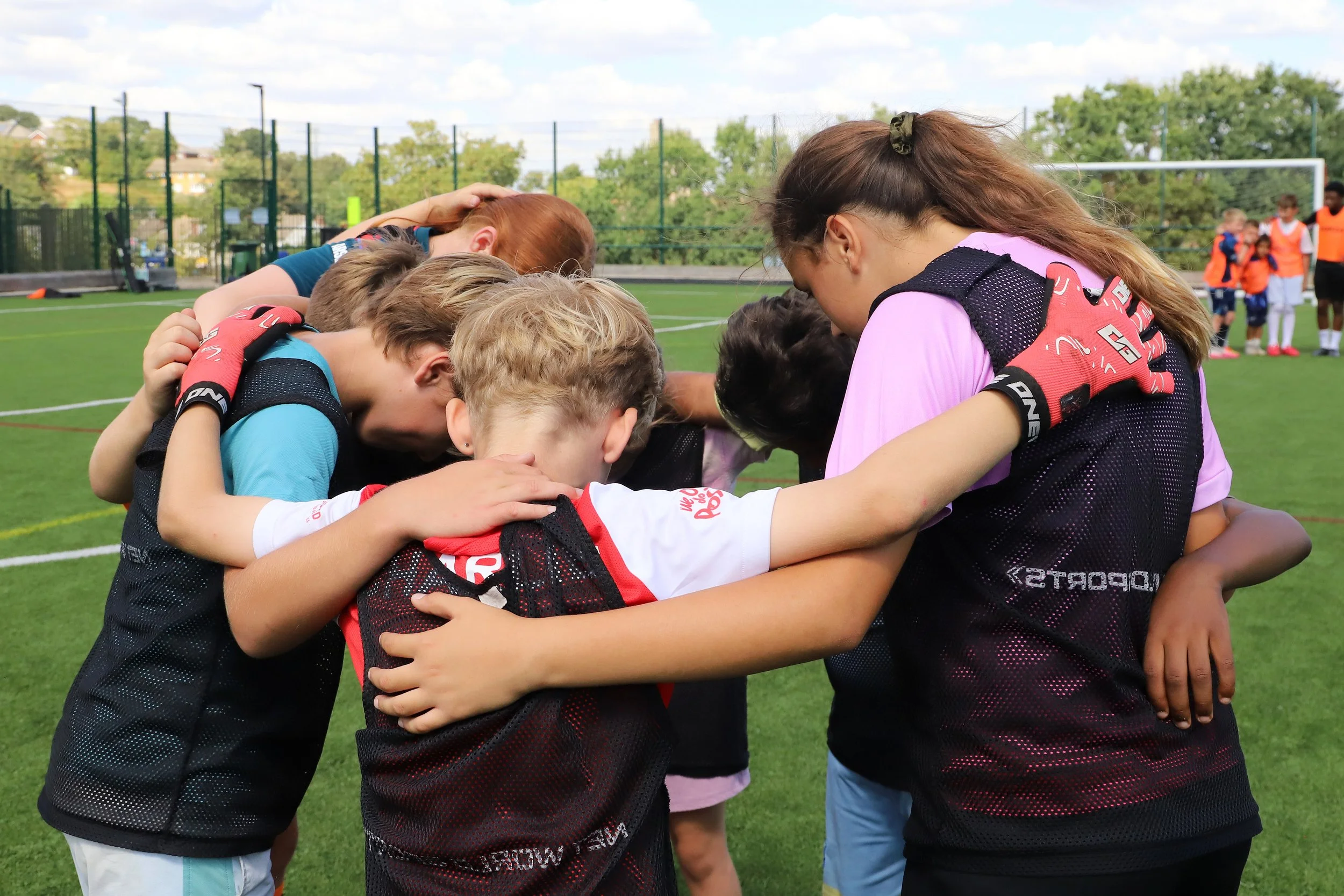 A group of young kids and a woman are huddled together with their heads bowed and arms around each other on a green outdoor sports field, likely before or after a game.