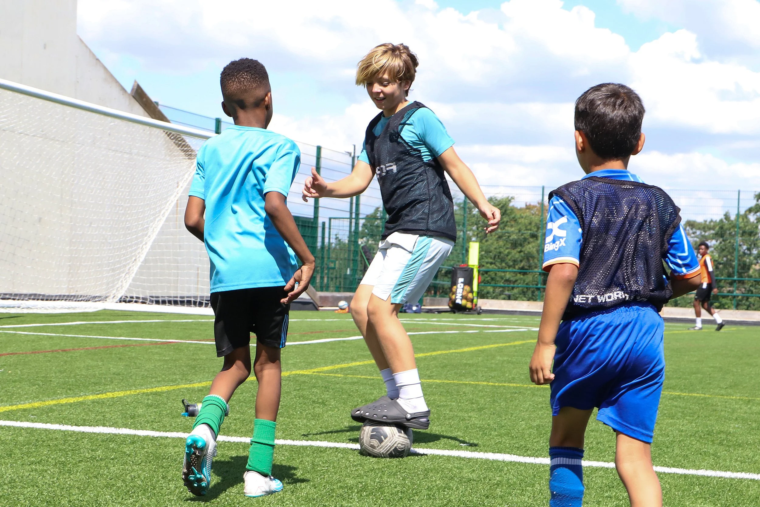 Children playing football on an outdoor field, three boys and a girl, with a goal net and a clear sky.