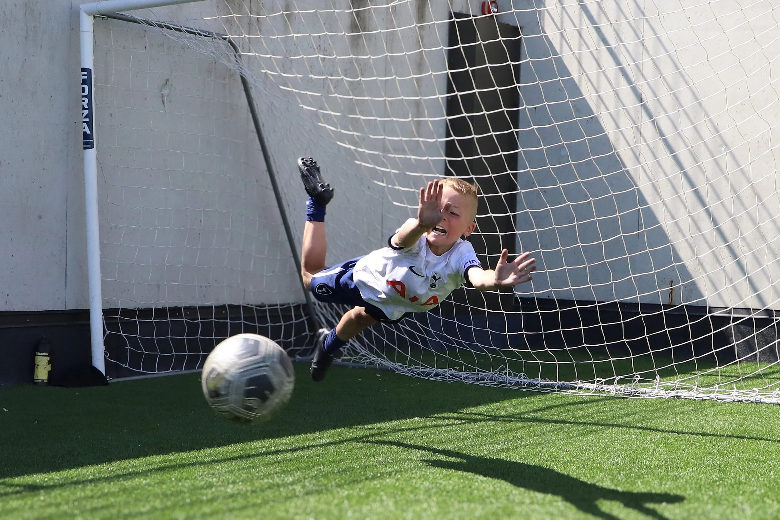 A young boy in a football uniform diving to save a soccer ball from going into the goal, with the net and goalpost visible in the background.