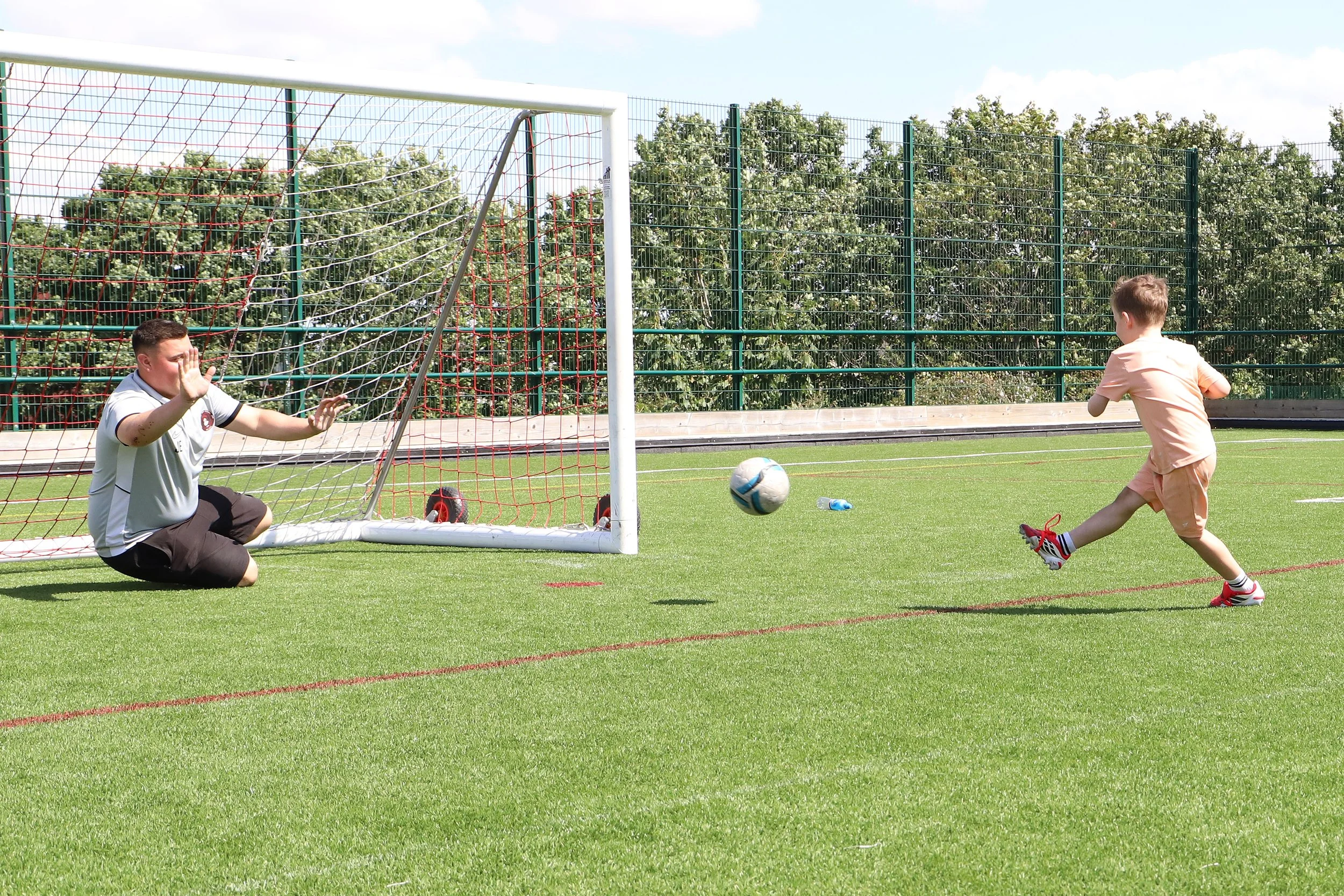 A young boy in a peach t-shirt and shorts is kicking a football ball towards a goal while a goalkeeper in a white shirt and dark shorts is getting ready to block it on a football field with a green fence and trees in the background.