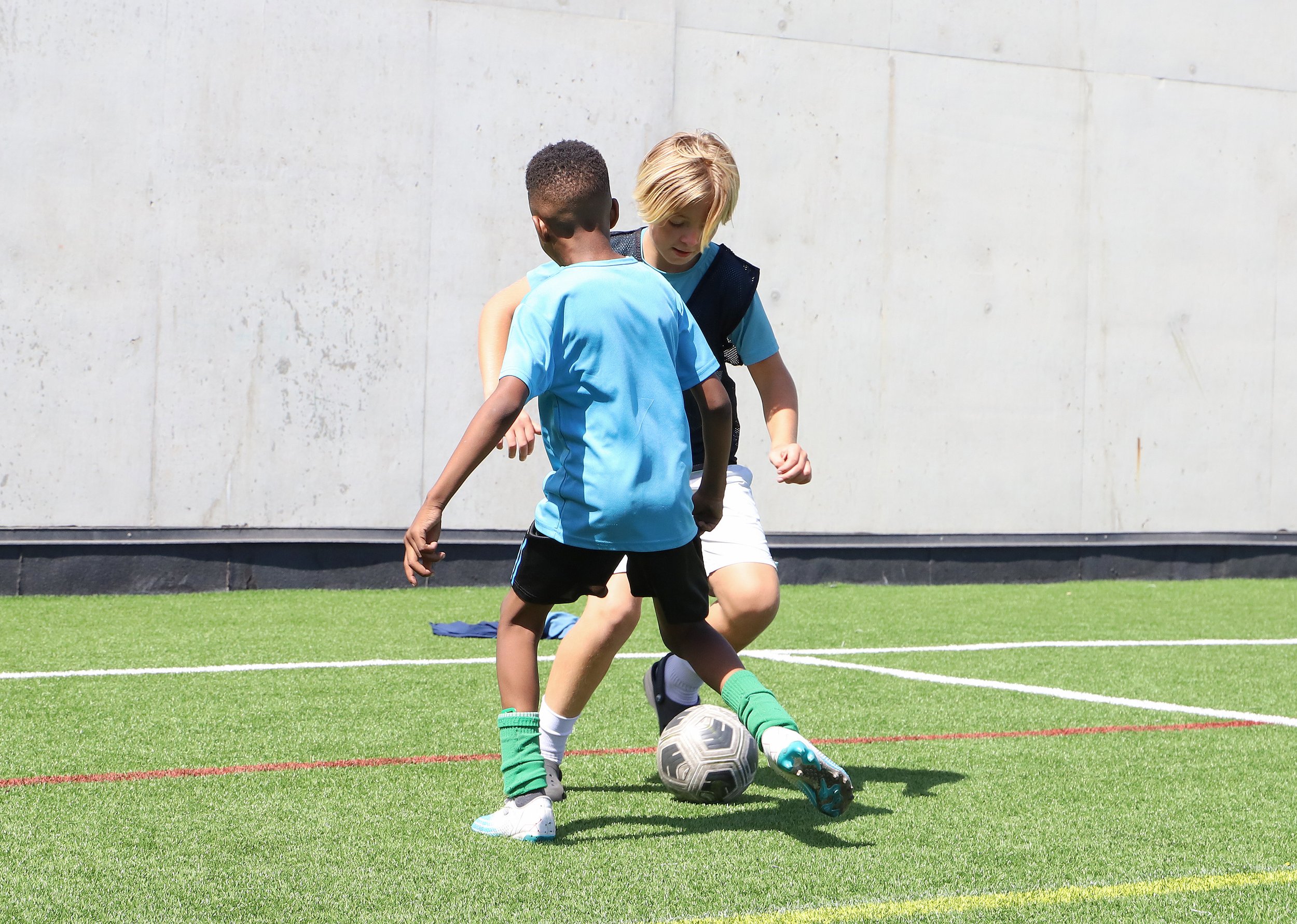 Two children playing football on a field, with one child in a blue shirt and shorts and the other with blonde hair in a darker shirt, both focused on the soccer ball.