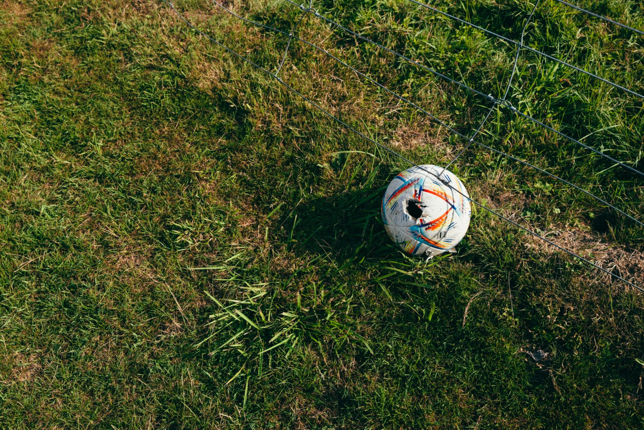 Un ballon de football blanc avec des éclaboussures de couleurs, posé sur de l'herbe, à côté d'une clôture en fil métallique