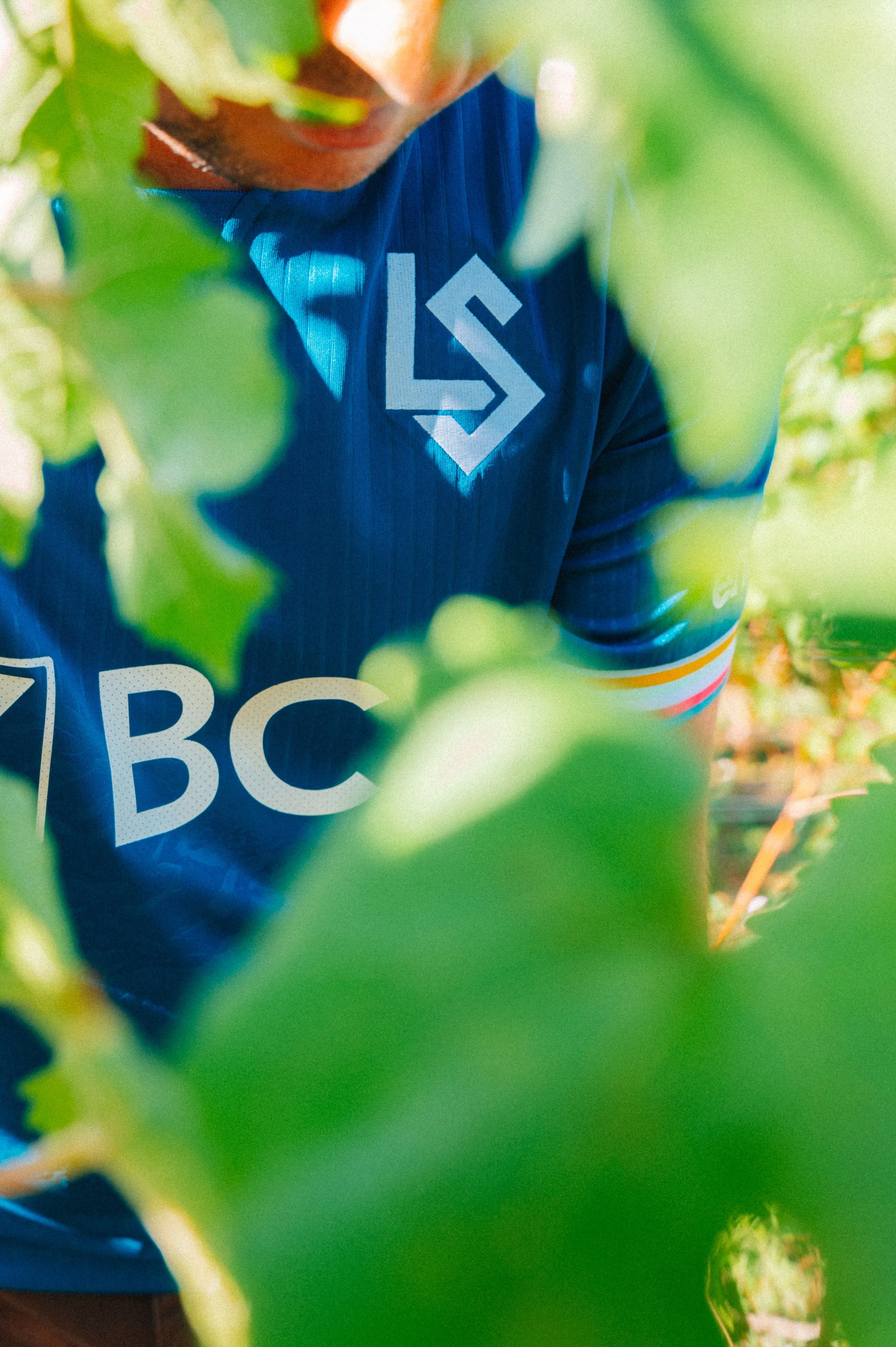 Premier plan d'une personne portant un t-shirt bleu avec un logo blanc, entourée de feuillage vert dans une serre ou un jardin.