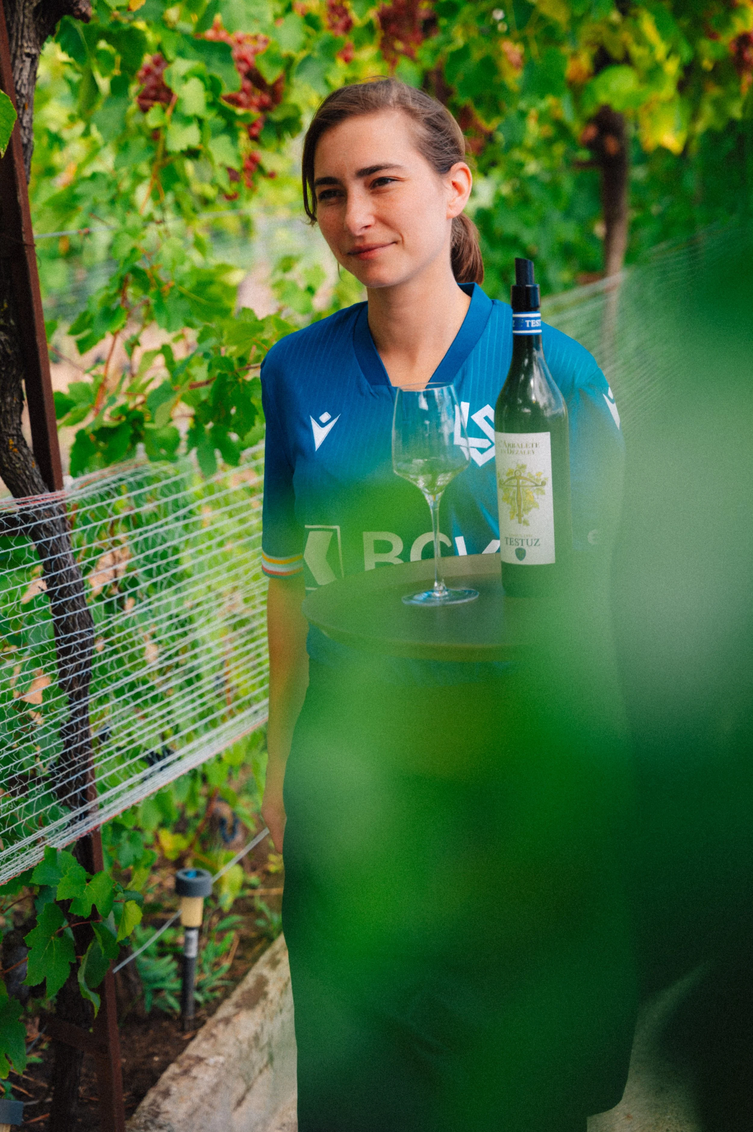 Une femme portant un maillot de sport bleu se trouve dans un vignoble, avec une bouteille de vin, un verre à vin vide et une bouteille de spray posés devant elle.