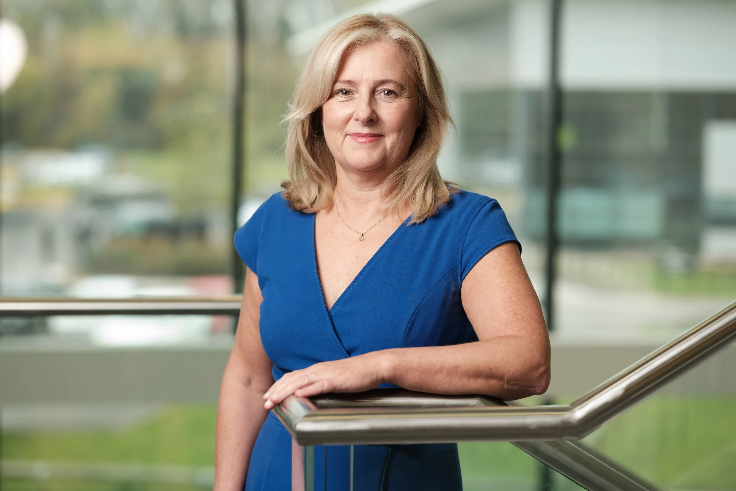 A woman with blonde hair wearing glasses and a light pink blouse, standing against a plain white wall.