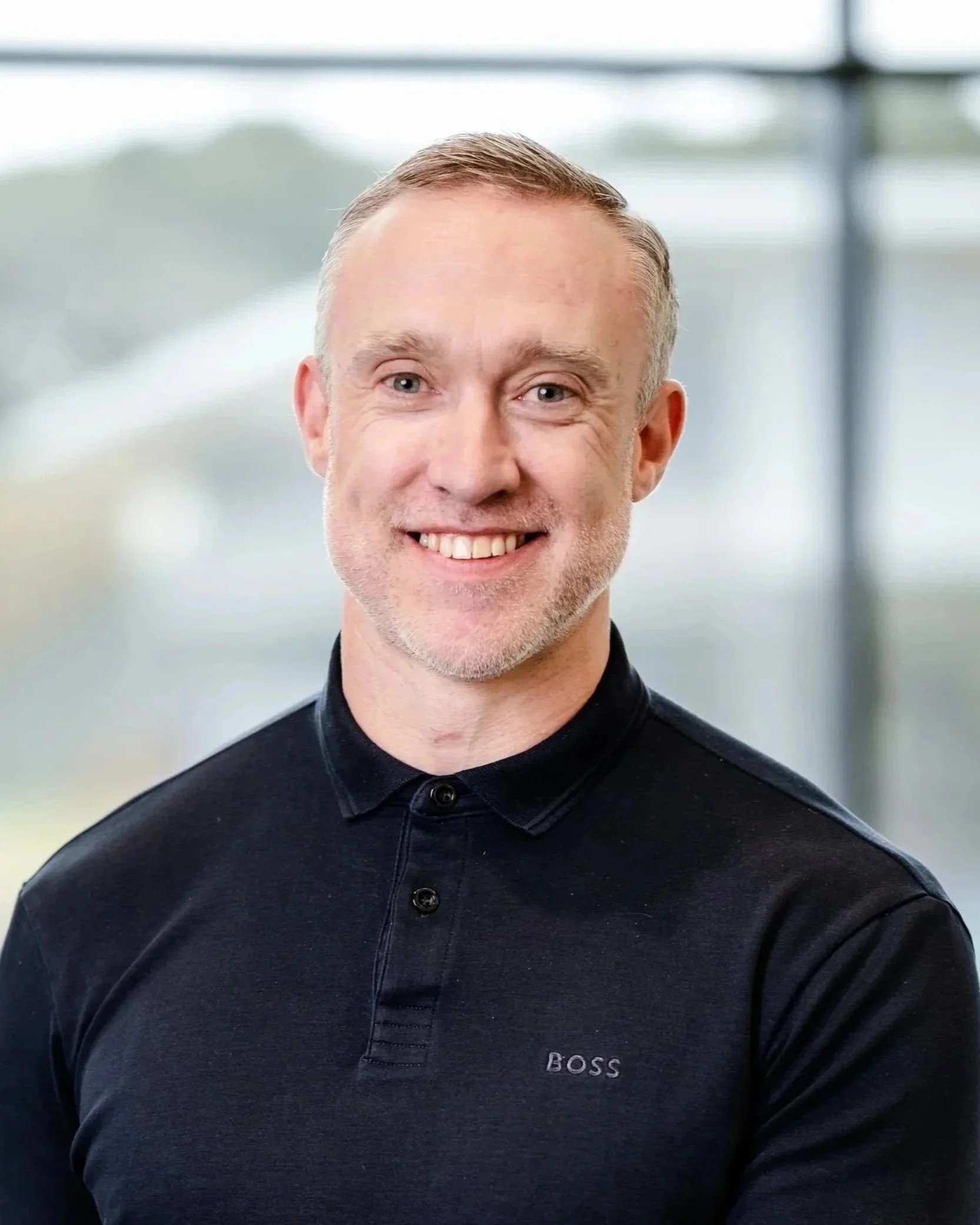 Headshot of a smiling man with short light hair, wearing a light blue collared shirt, against a blue background.