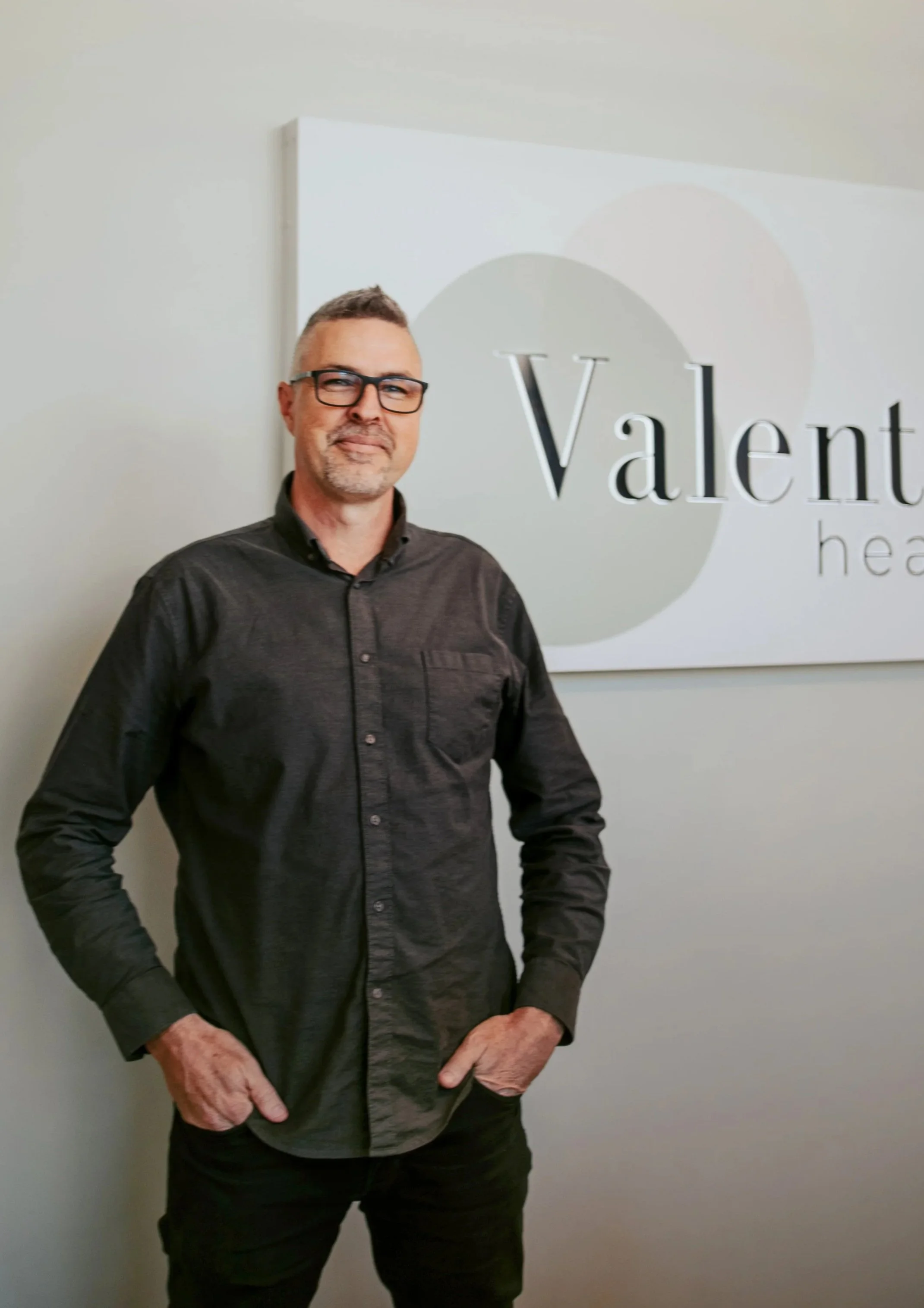 A middle-aged man with glasses, gray hair, and a beard, wearing a dark button-up shirt, standing in front of a white wall with a sign that reads 'Valent' and some smaller, unclear text.