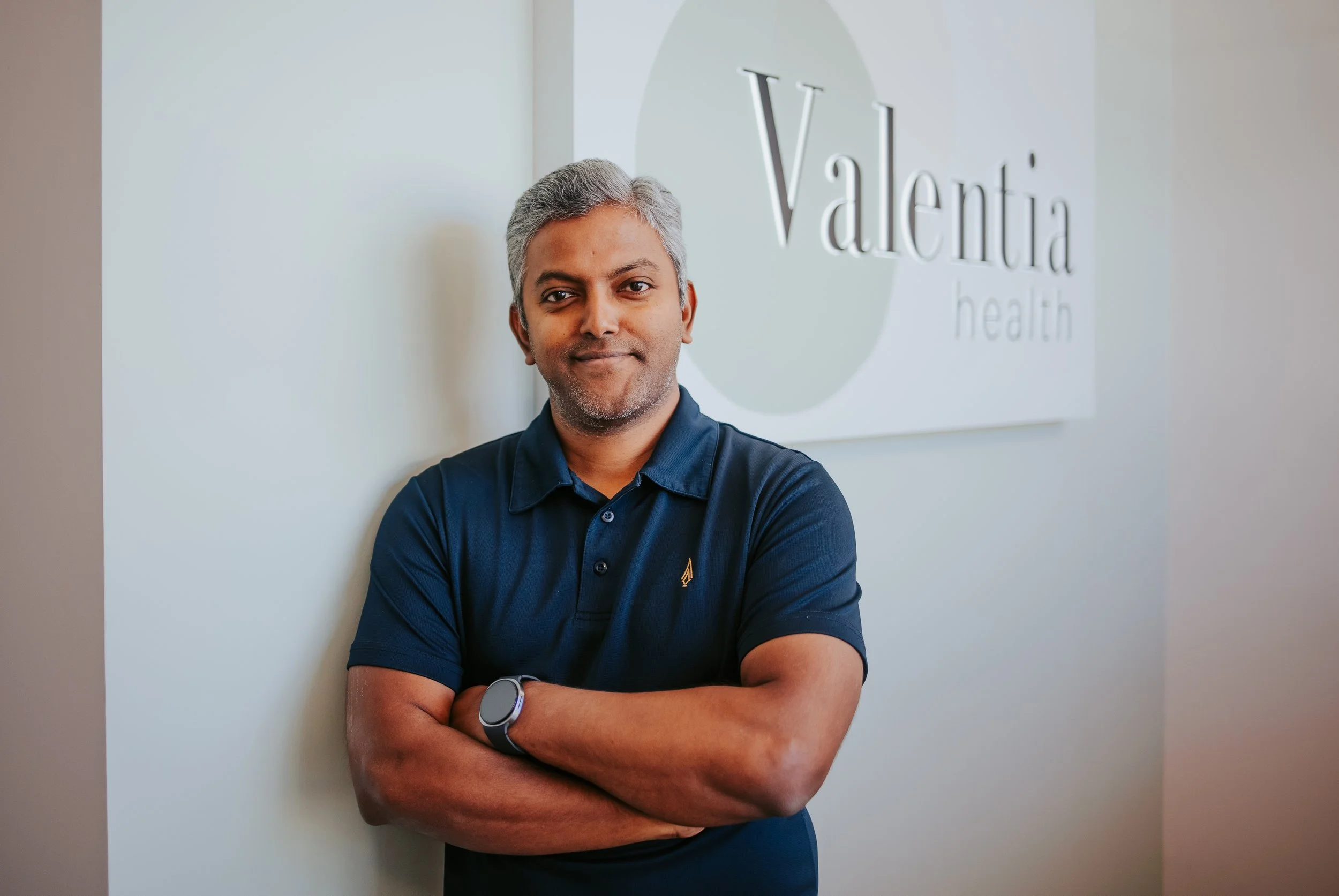 A man with gray hair and a mustache standing with arms crossed, wearing a navy blue polo shirt and a smartwatch, in front of a white wall with a sign that reads 'Valentia health'.