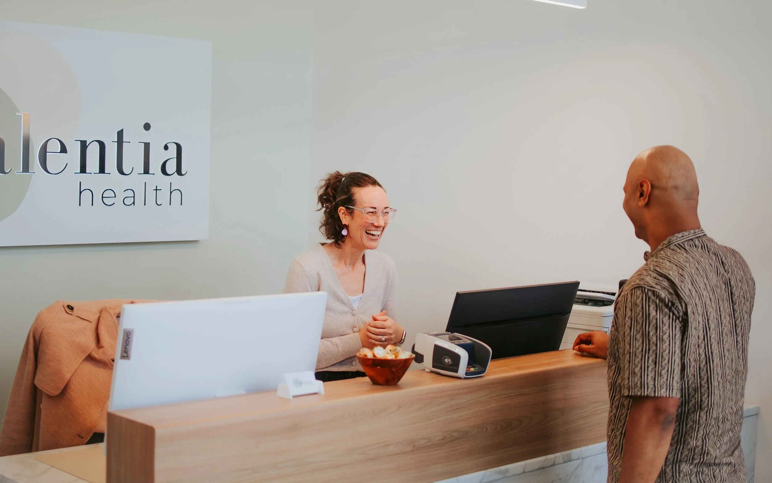 A woman with curly hair and glasses sitting at a front desk, smiling while speaking with a man with a shaved head and patterned shirt in an office with a sign that reads 'Valentia Health' on the wall.