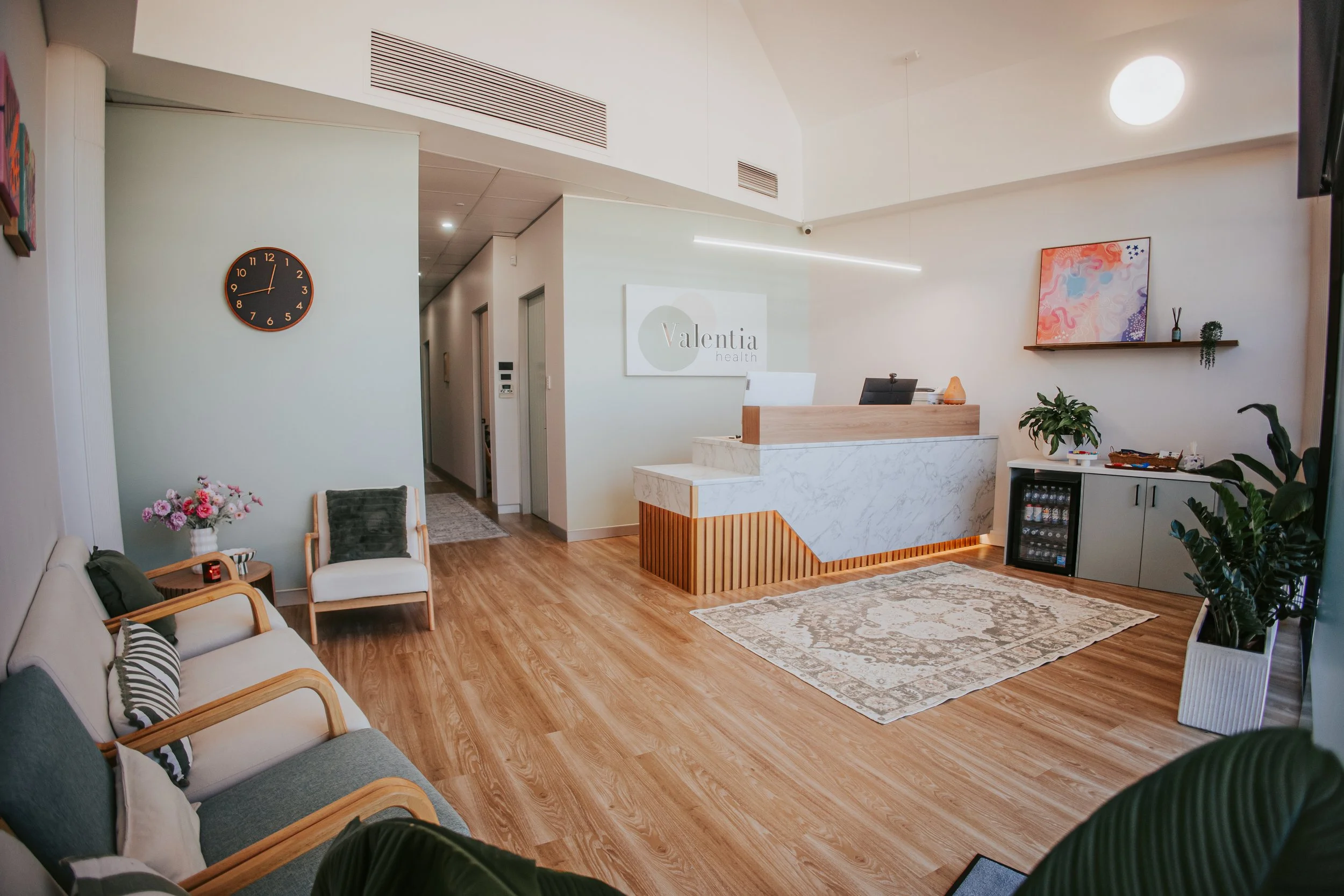The reception area of Valentina Health featuring a front desk with marble and wood design, a sign with the company logo on the wall, seating with white and gray chairs, multiple plants, colorful artwork, a large wall clock, and a cozy rug on wooden flooring.
