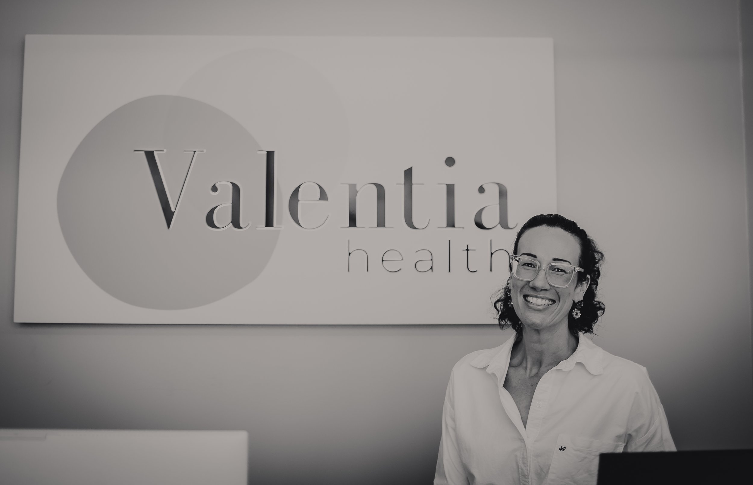 A smiling woman with glasses and earrings standing in front of a banner that reads 'Valentia health' with a geometric design. The woman is wearing a white shirt and appears to be in an office or clinic environment.