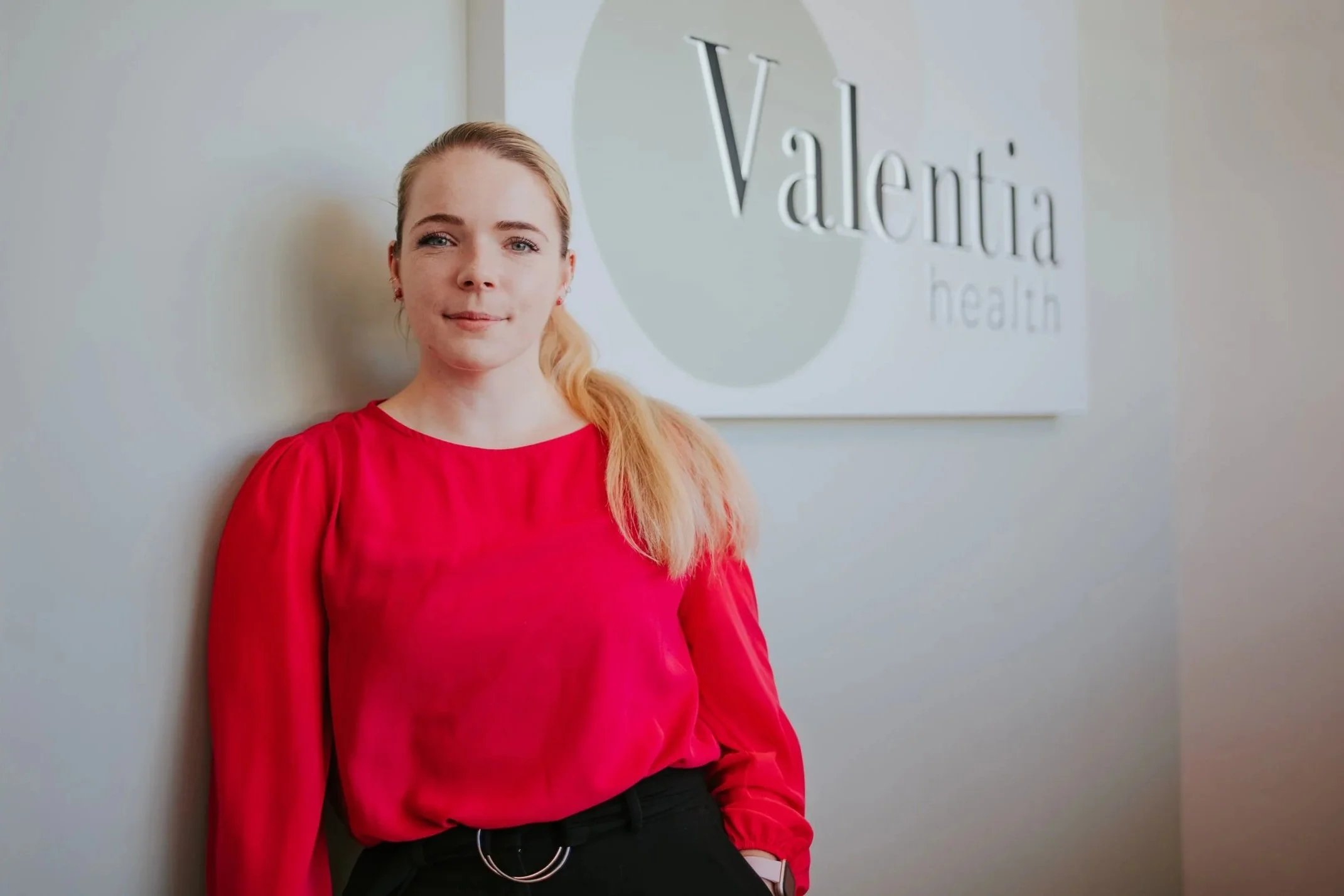 A woman with blonde hair wearing a red blouse, standing against a wall with a logo that reads 'Valentia health'.