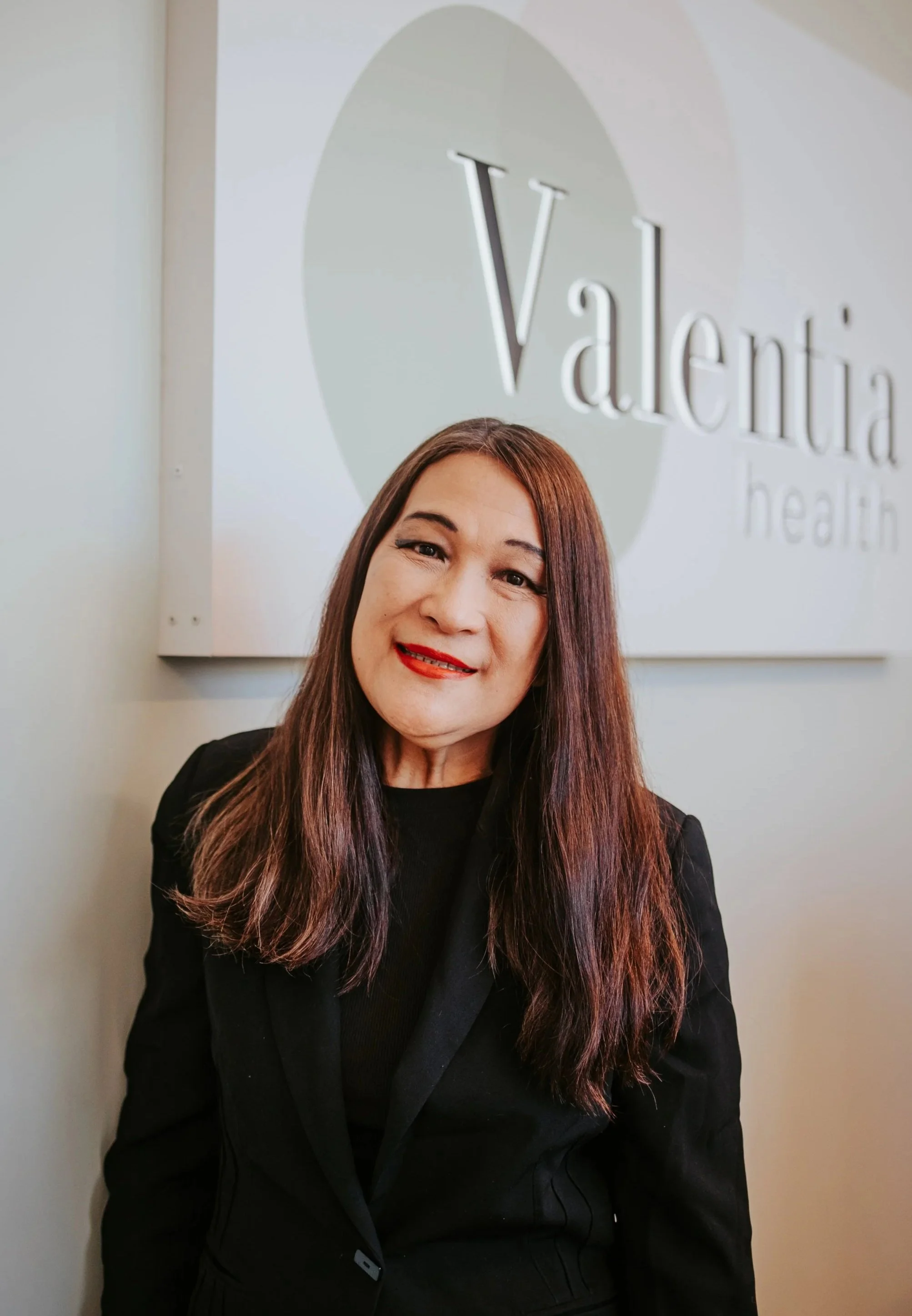 A woman with long brown hair and red lipstick smiling in front of a wall with a sign that reads 'Valentia health'.