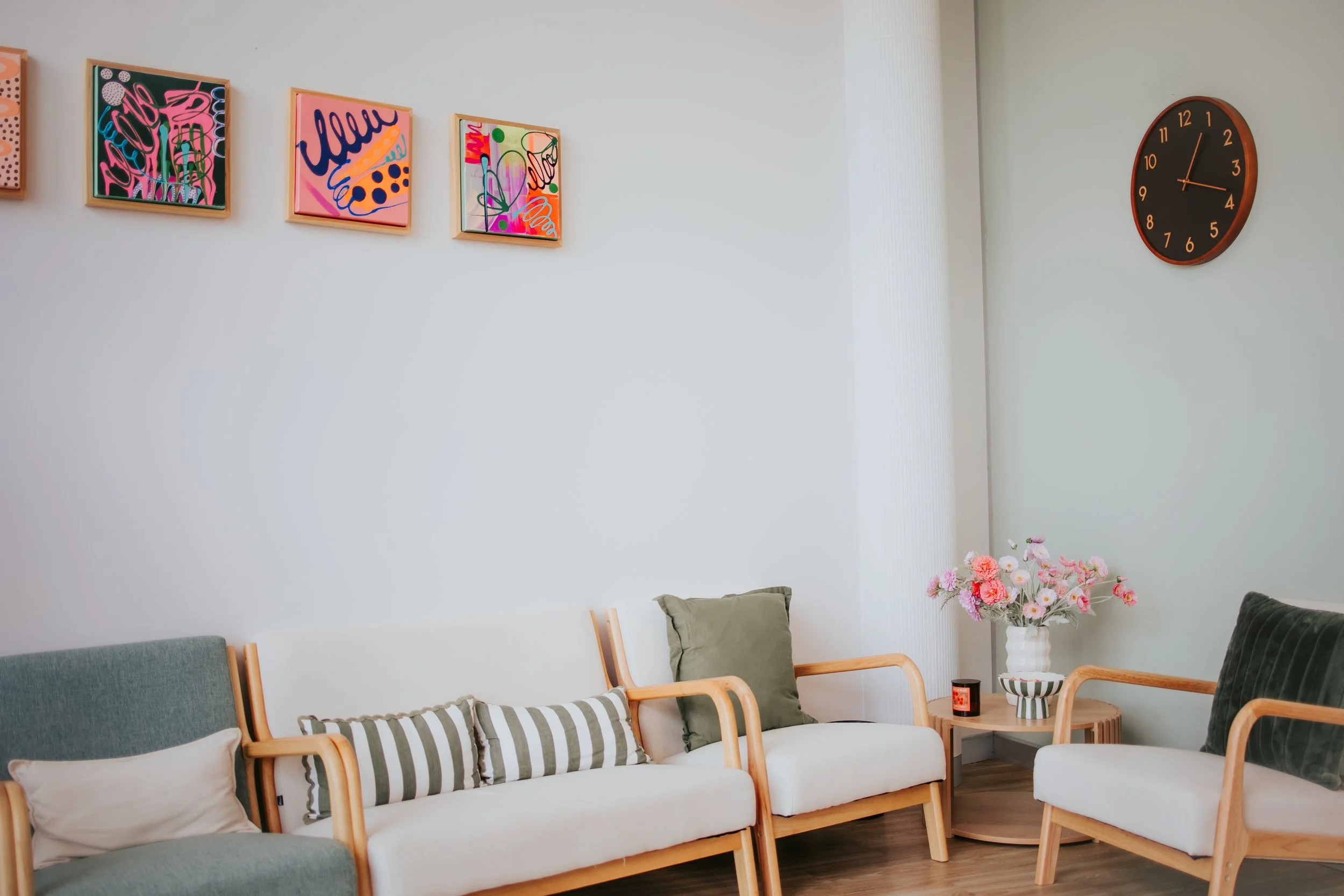 A living room with a white wall decorated with colorful abstract framed artworks. A black wall clock shows the time as 1:13. Furniture includes a seating area with green and white cushions and a small wooden side table holding a white vase with pink and white flowers, a striped bowl, and candles.