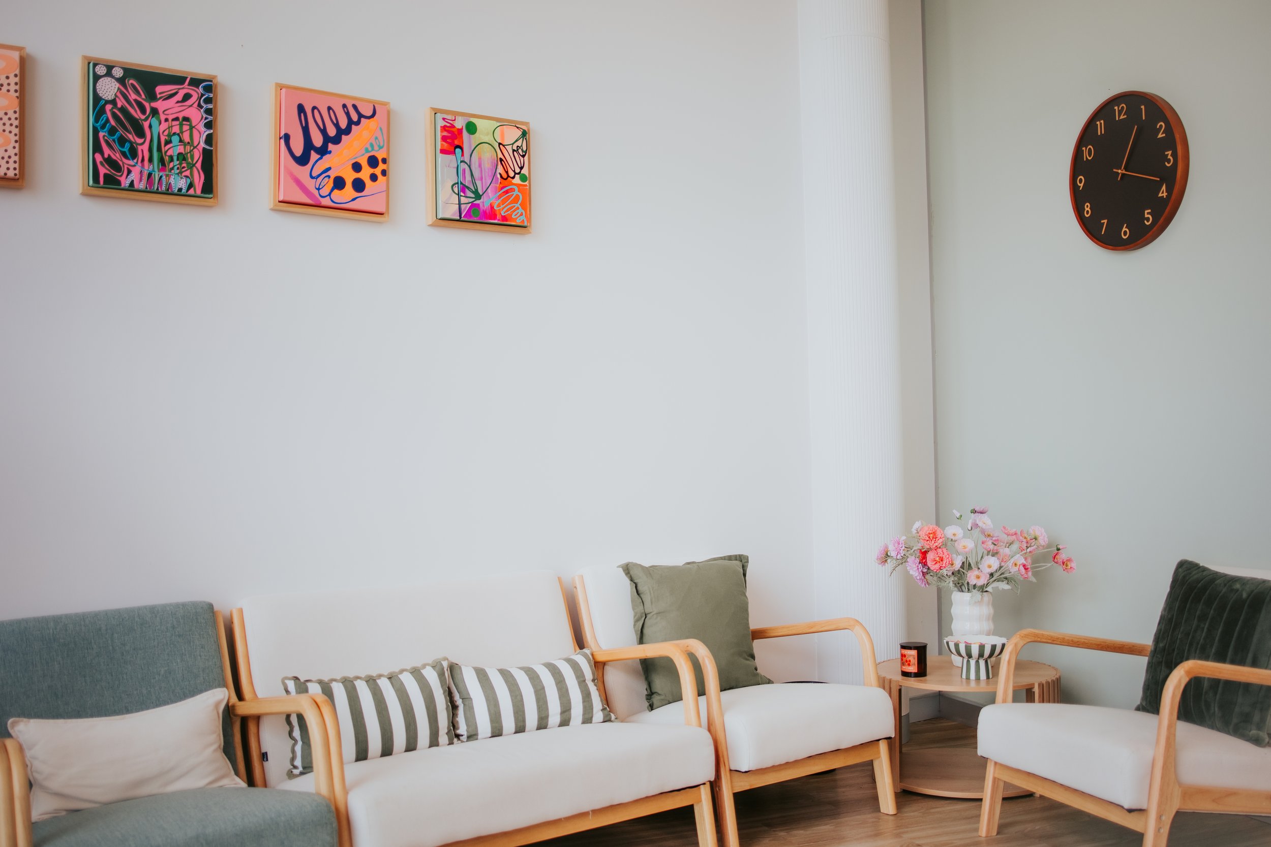 A living room with a white wall decorated with colorful abstract framed artworks. A black wall clock shows the time as 1:13. Furniture includes a seating area with green and white cushions and a small wooden side table holding a white vase with pink and white flowers, a striped bowl, and candles.