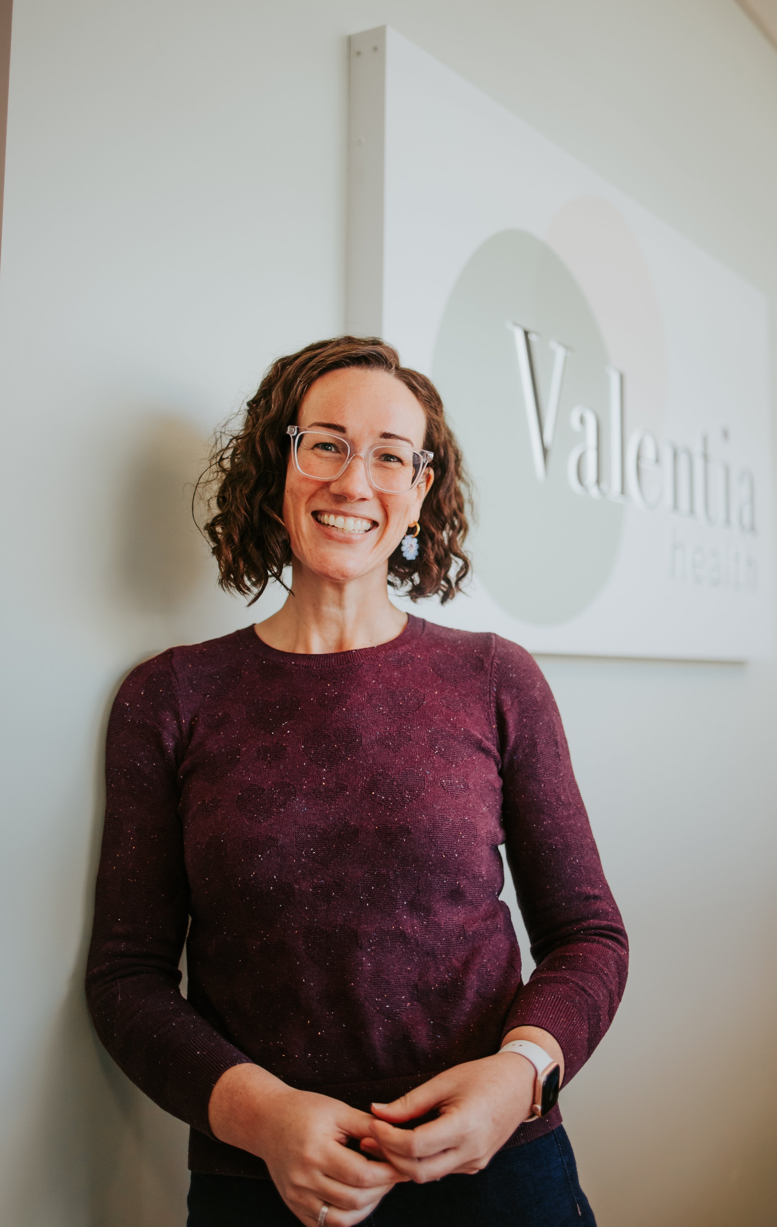 A smiling woman with short curly brown hair, glasses, and a floral earring, wearing a maroon glittery long-sleeve shirt, standing in front of a wall with a logo that reads 'Valentia'.