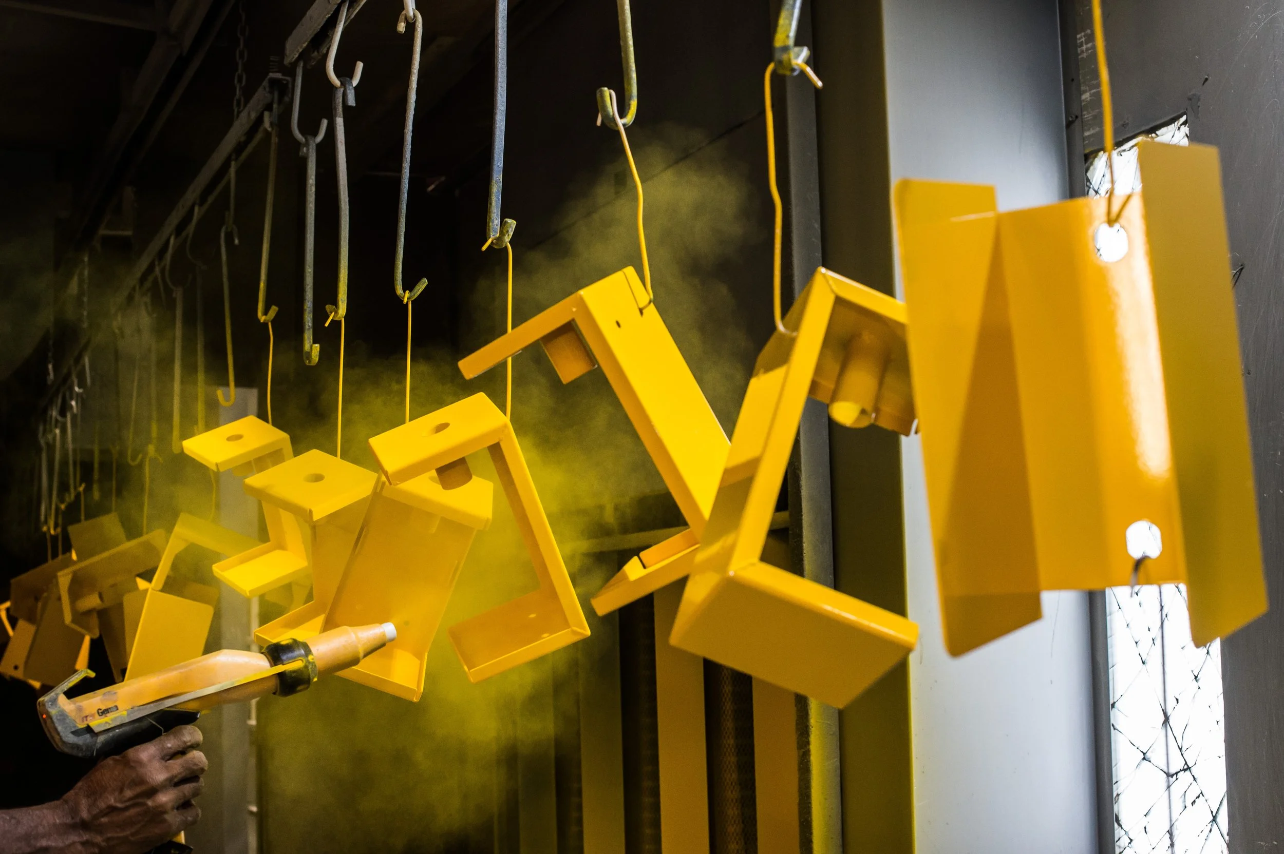 Yellow painted metal objects hanging in a workshop, some being sprayed with yellow paint, with a person using a spray gun to paint them.