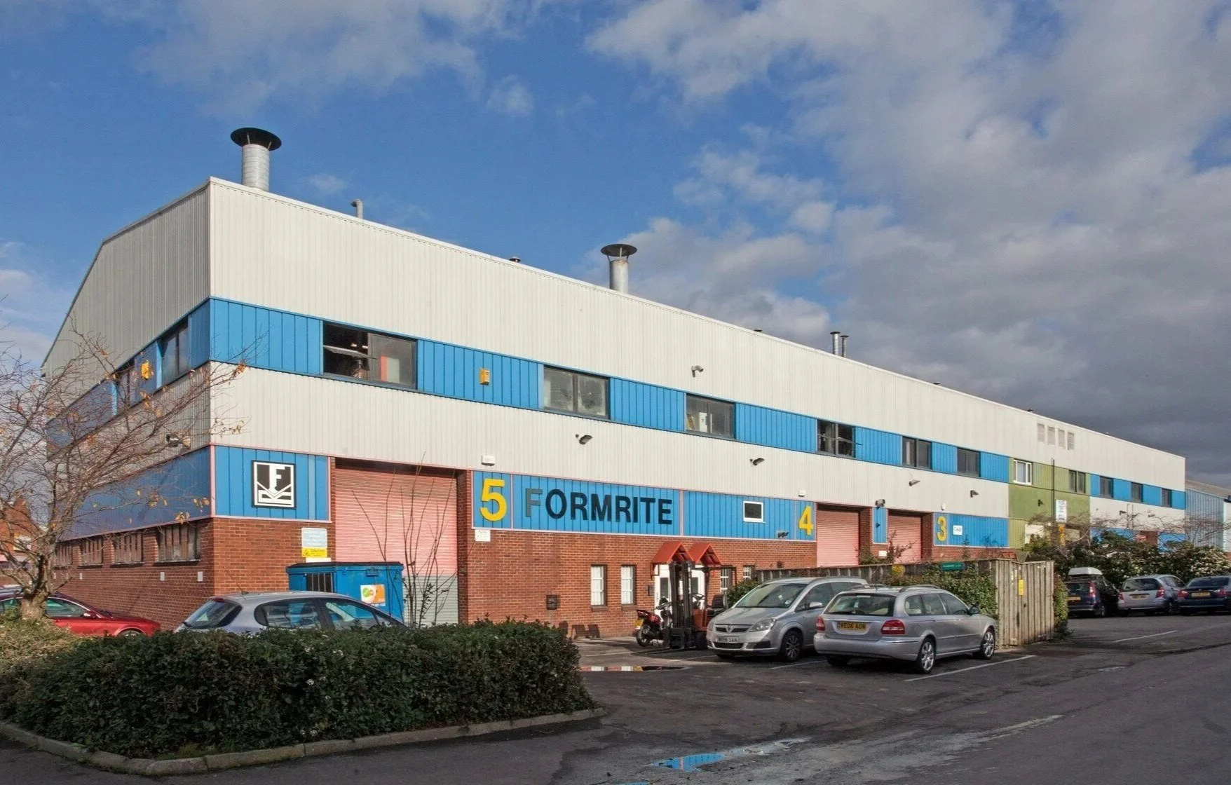 A large industrial warehouse building with a brick and blue metal exterior, multiple loading docks, and a parking area in front. The building has signs indicating sections 3, 4, and 5, and a company logo reading Formrite.