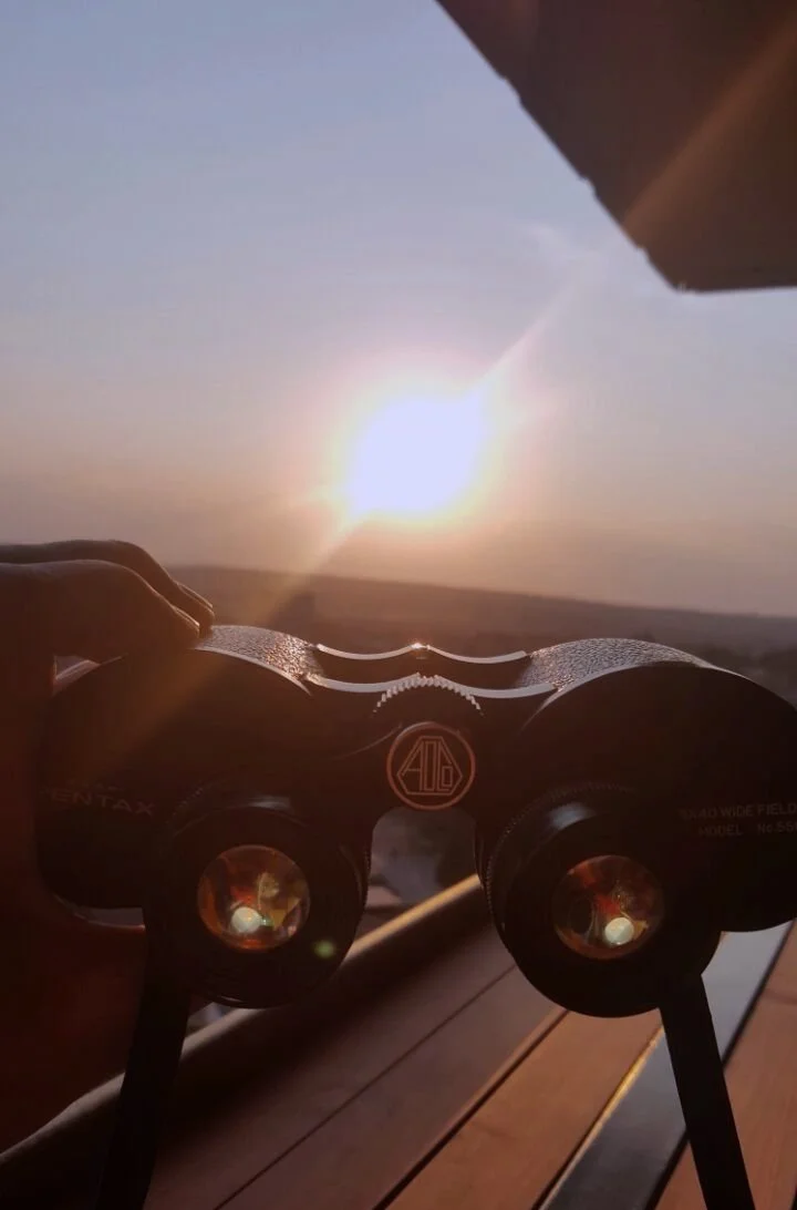 Person holding binoculars to watch the sun setting or rising over a flat landscape.