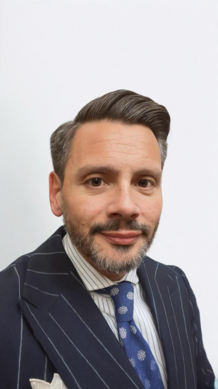 A man with a beard and short hair wearing a checkered blazer, white shirt, pink tie, and a white pocket square, standing against a white background.