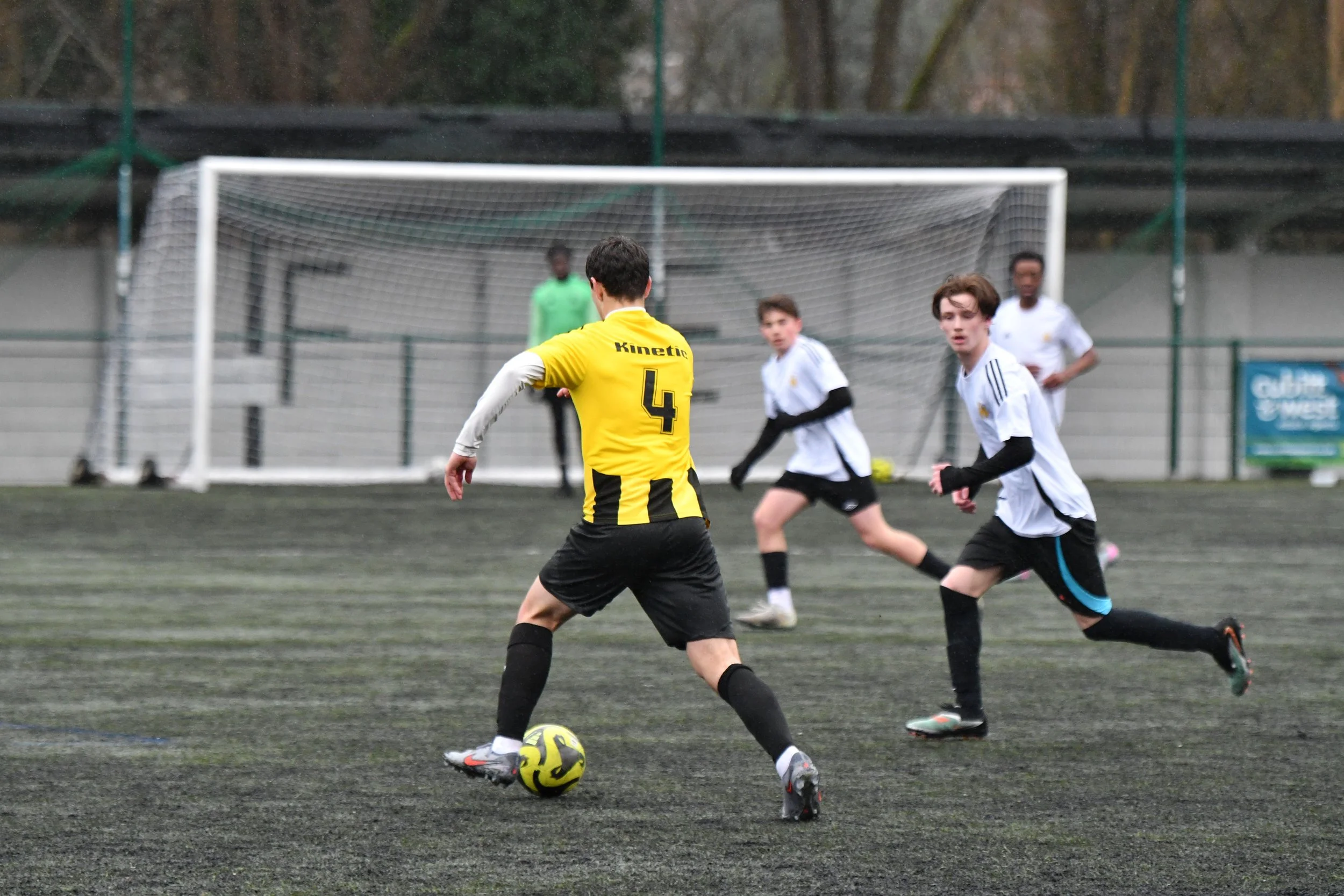 Young soccer players during a match on an outdoor field, focusing on one player in a yellow jersey with the number 4, about to kick a yellow and black soccer ball, with a goalpost and other players in white jerseys in the background.