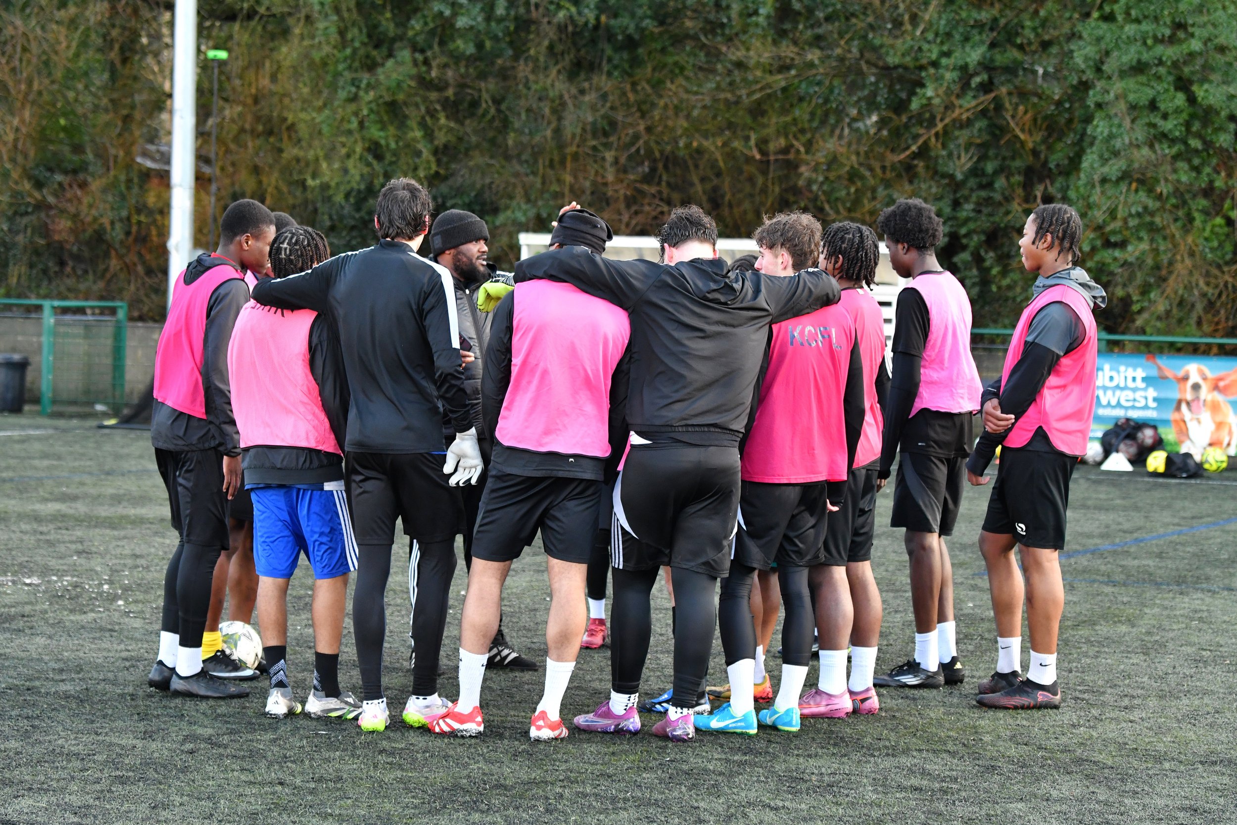 A group of soccer players and coaches huddling together on a soccer field during a practice or game, wearing pink bibs and athletic gear, with trees and a sign in the background.