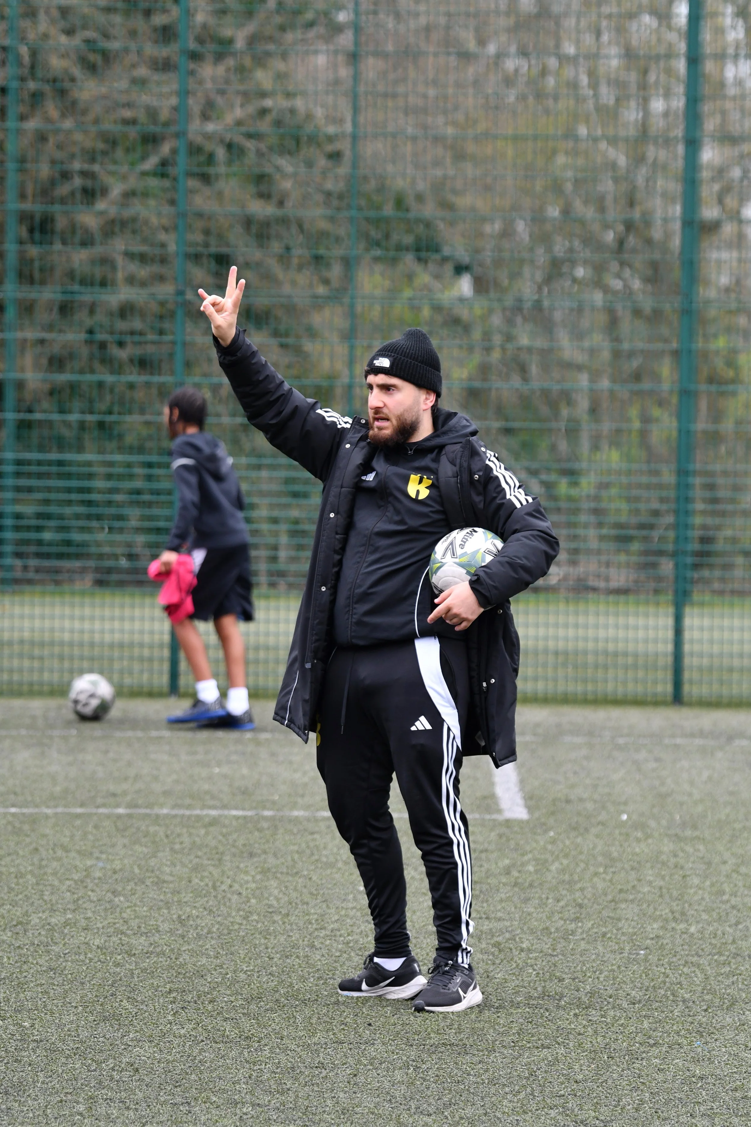 A man in black sportswear walking on a synthetic outdoor football field with a green fence and trees in the background.