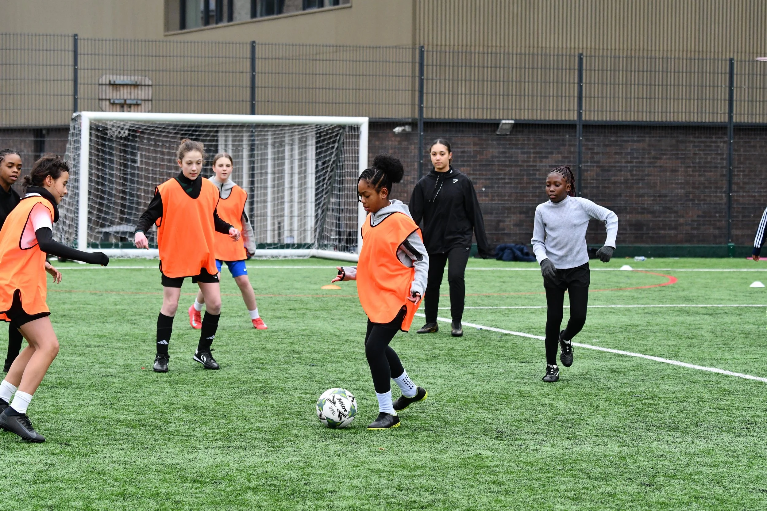 Group of young girls playing soccer on an outdoor field, some wearing orange practice vests, with a goal net and a building in the background.