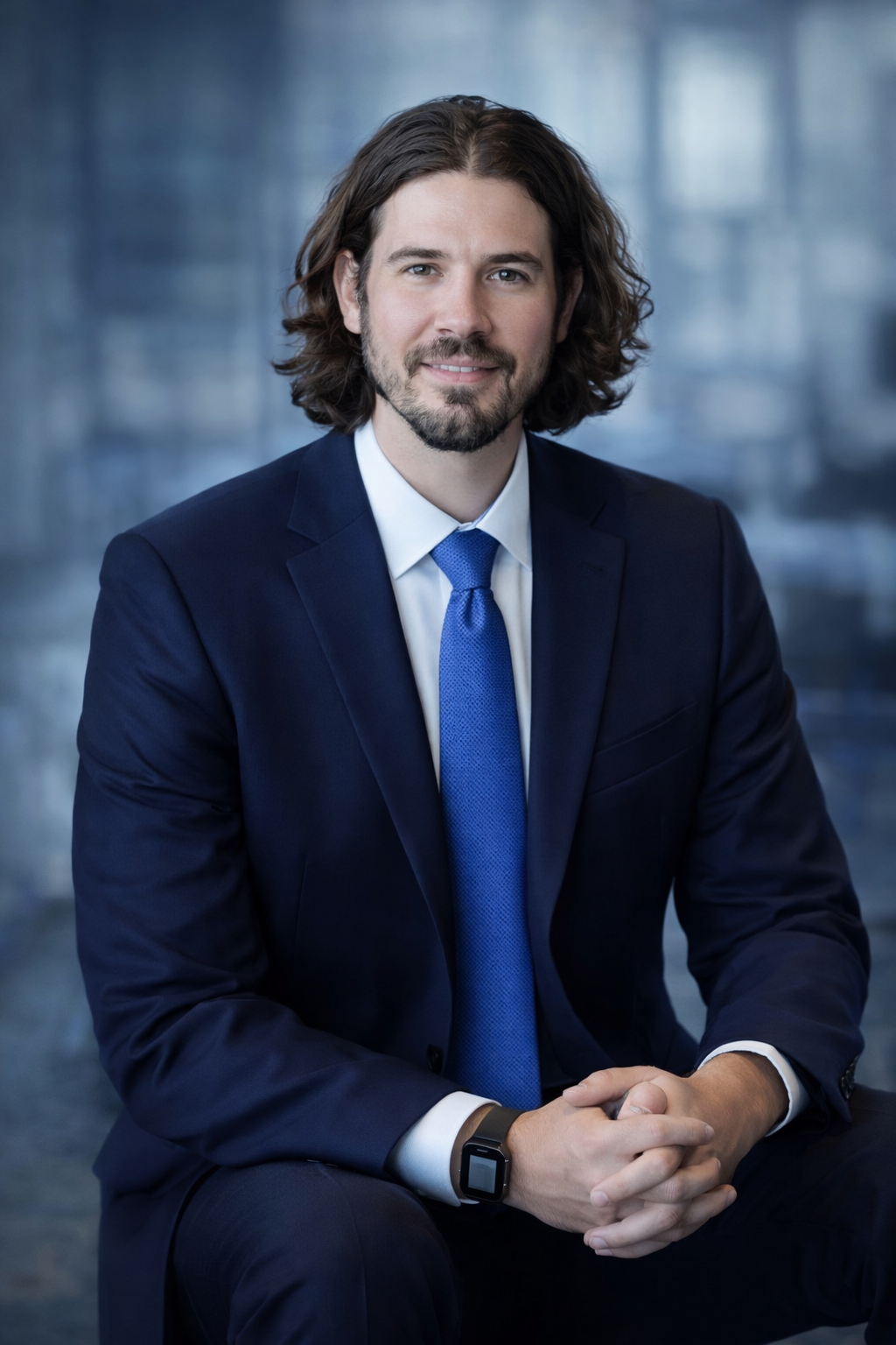 A man with shoulder-length brown hair and a beard, wearing a navy blue suit, white shirt, and blue tie, sitting with hands clasped in front of him, smiling at the camera in a professional setting.