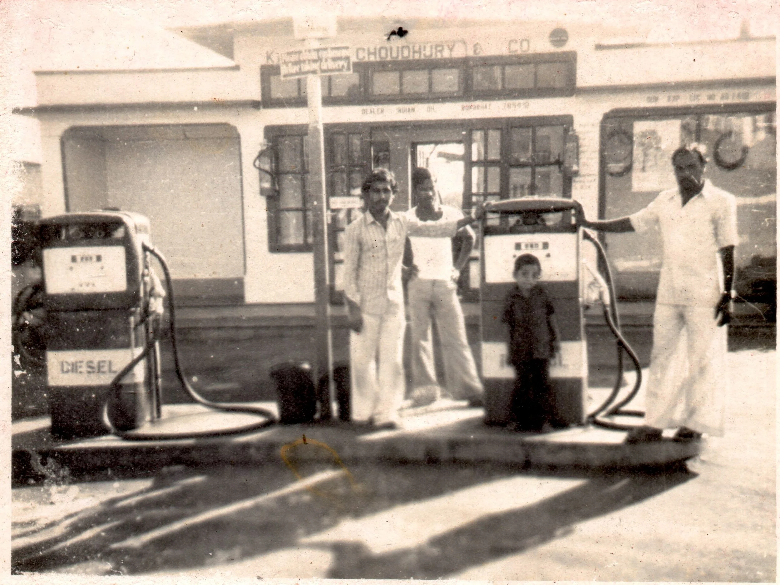 Black and white photo of three men and a boy standing at a vintage fuel station with diesel pumps, outside a building with signs, likely from the mid-20th century.