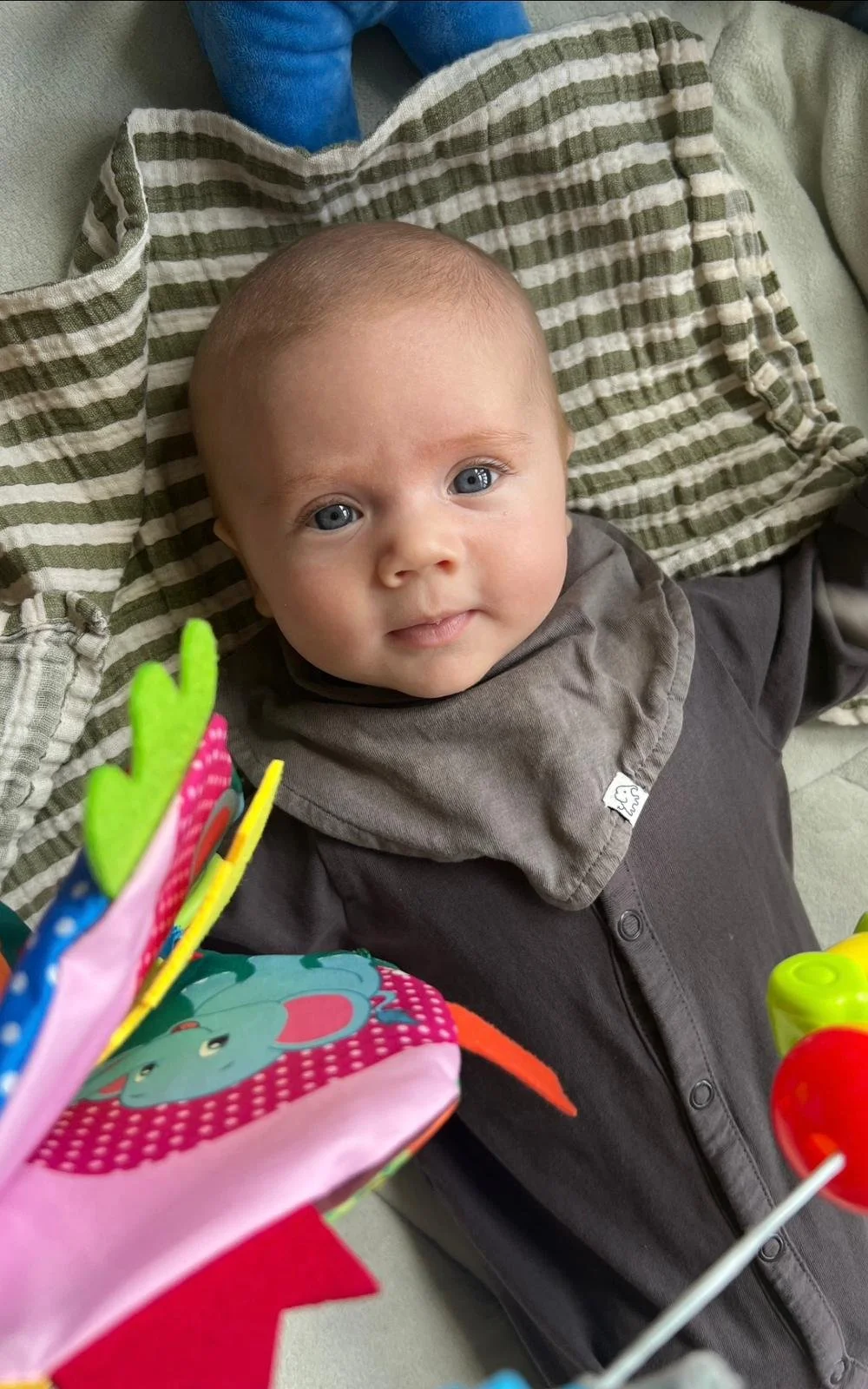 A baby with blue eyes and light hair lying on a blanket, gazing at the camera, with colorful toys partially visible in the foreground.