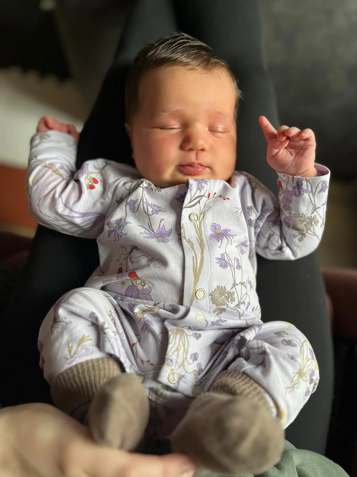 A sleeping baby dressed in a floral-patterned onesie, lying on a person's lap with eyes closed and a peaceful expression.