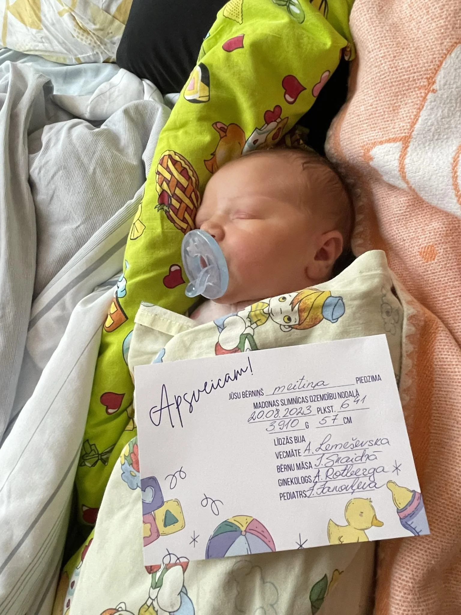 A newborn baby with a pacifier sleeping on a bed, dressed in a colorful cartoon-themed outfit, and holding a birth announcement sign with handwritten details.