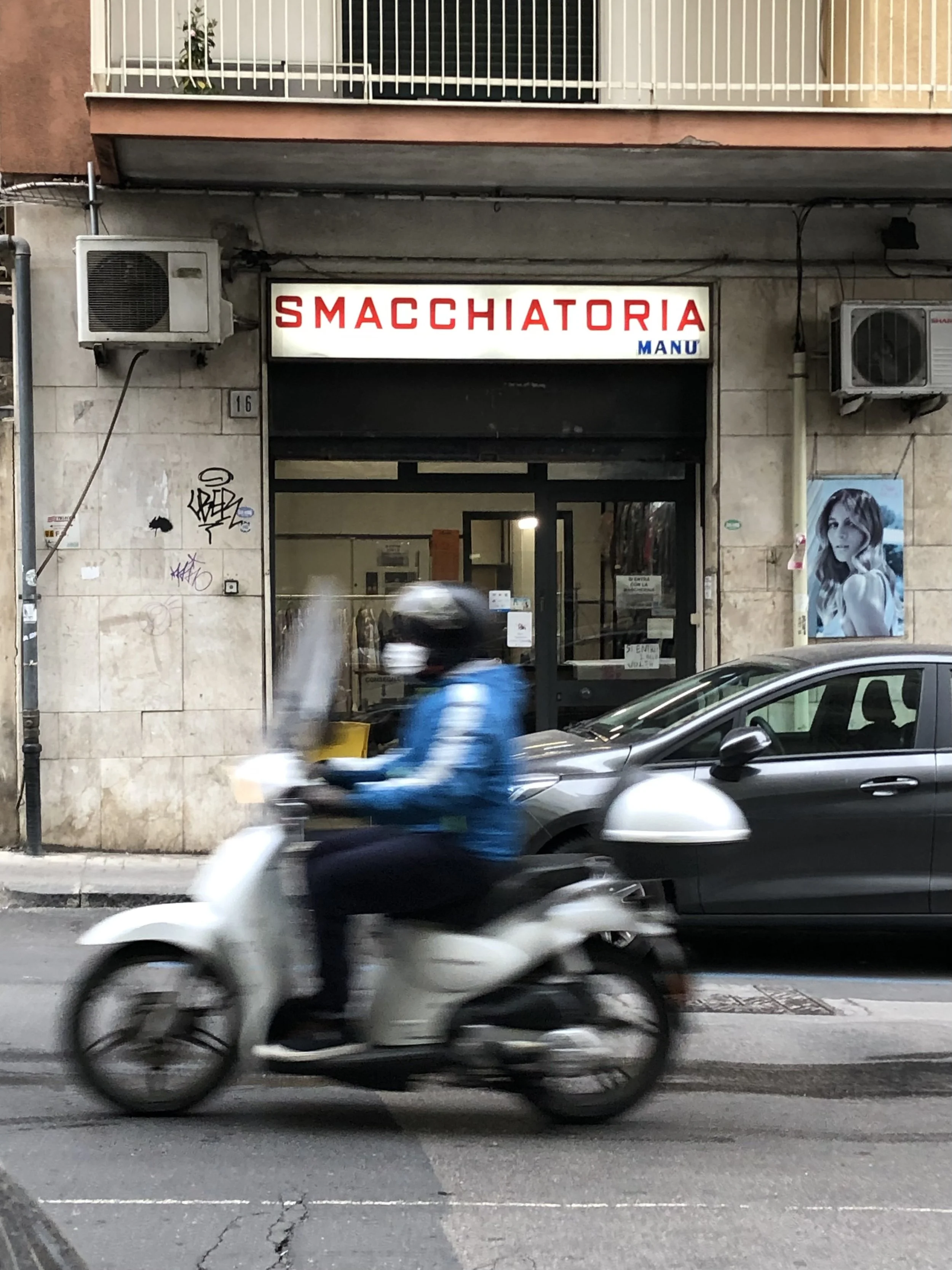 A motorcycle driving by a business on a typical street in Catania, Sicily.