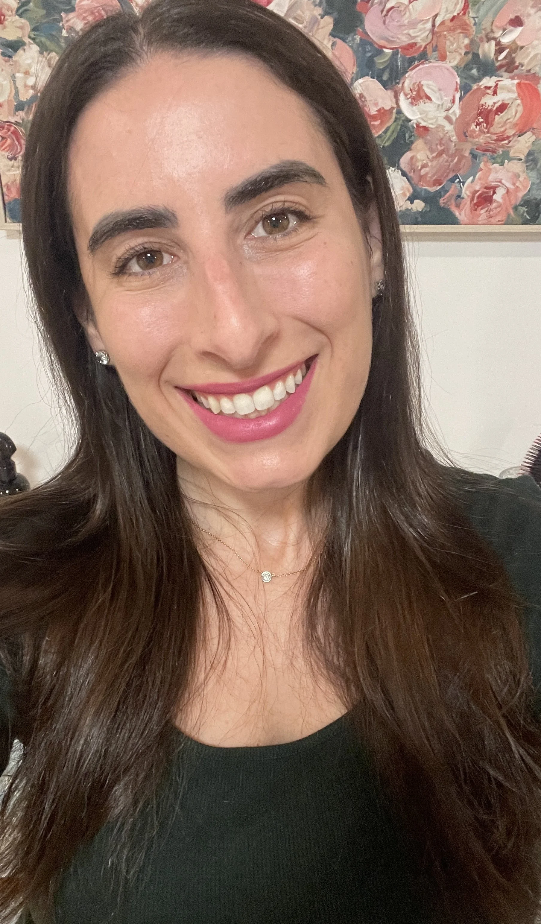 Close-up of a woman with long brown hair smiling, wearing a black top and a delicate necklace, with a floral painting in the background.