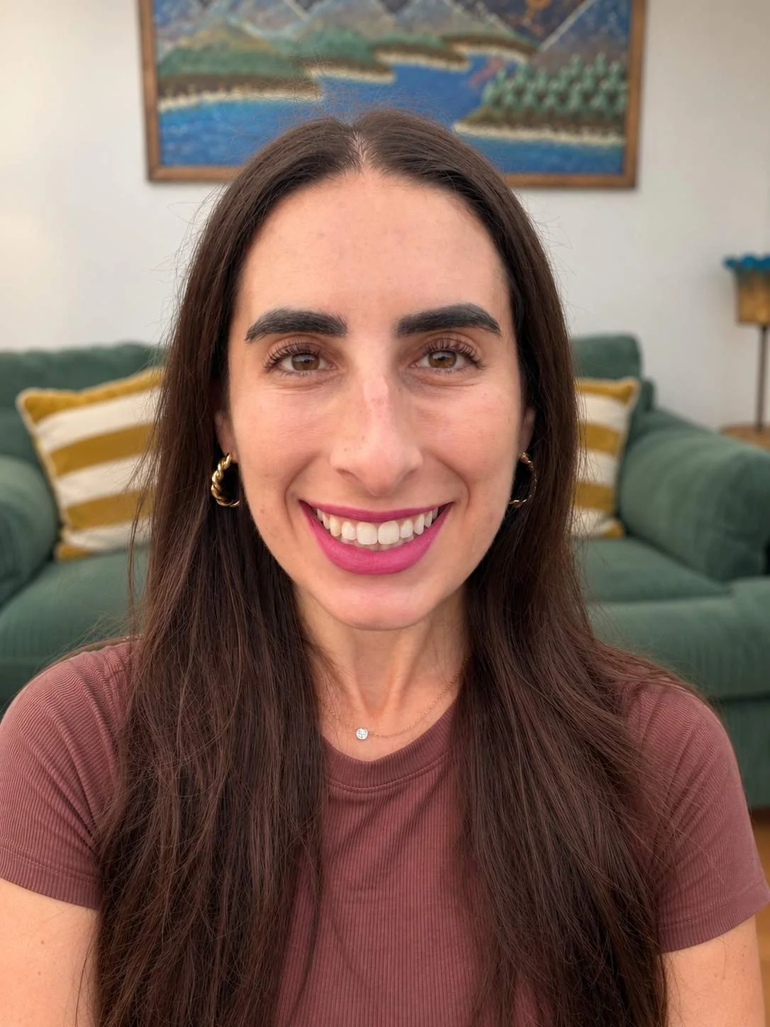 Lane, a woman with long brown hair, smiling, standing in front of couch with colorful mountain and lake artwork behind it