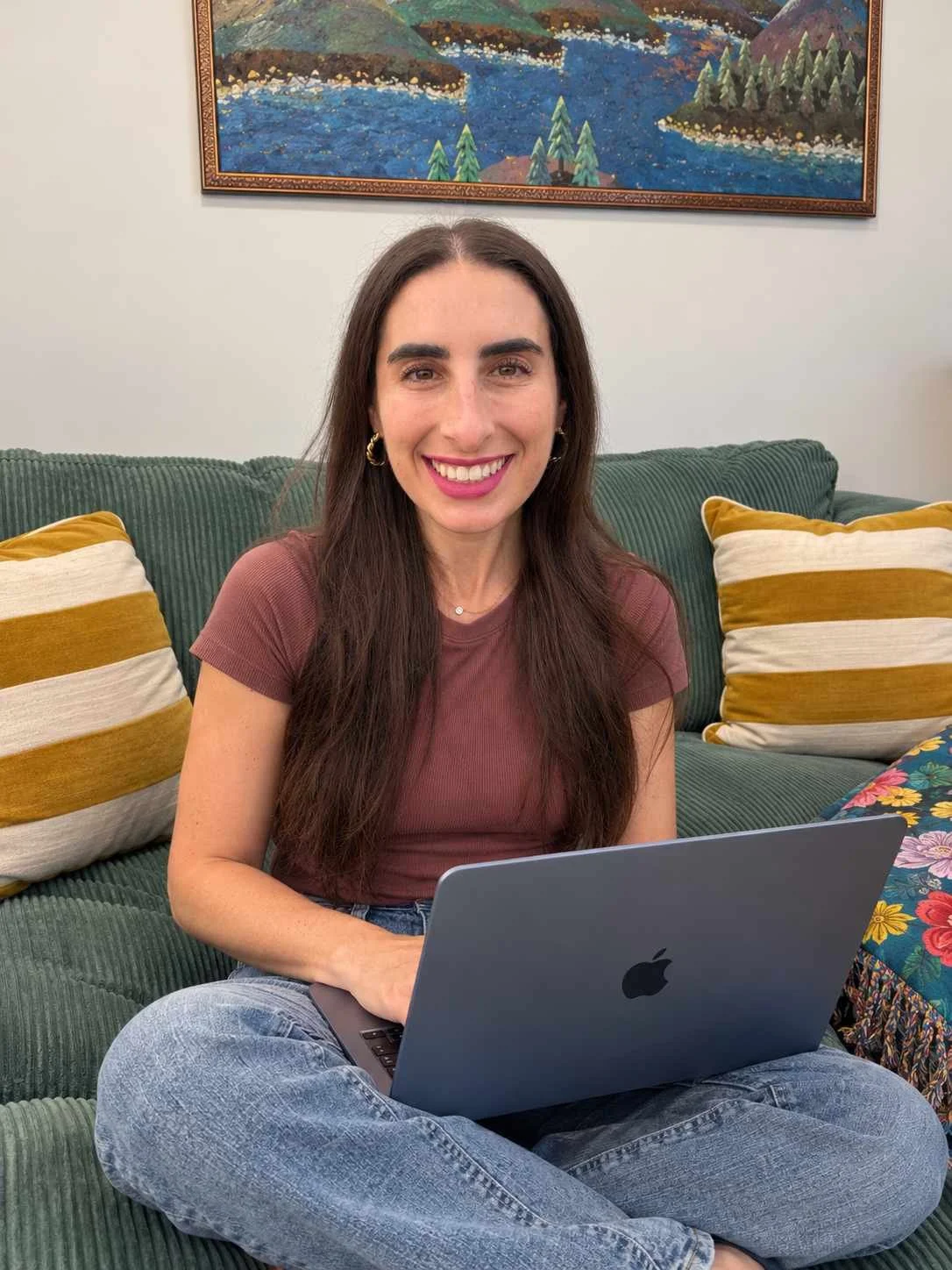 Lane, a woman with long brown hair, smiling, sitting on couch with laptop on lap in front of colorful mountain and lake artwork