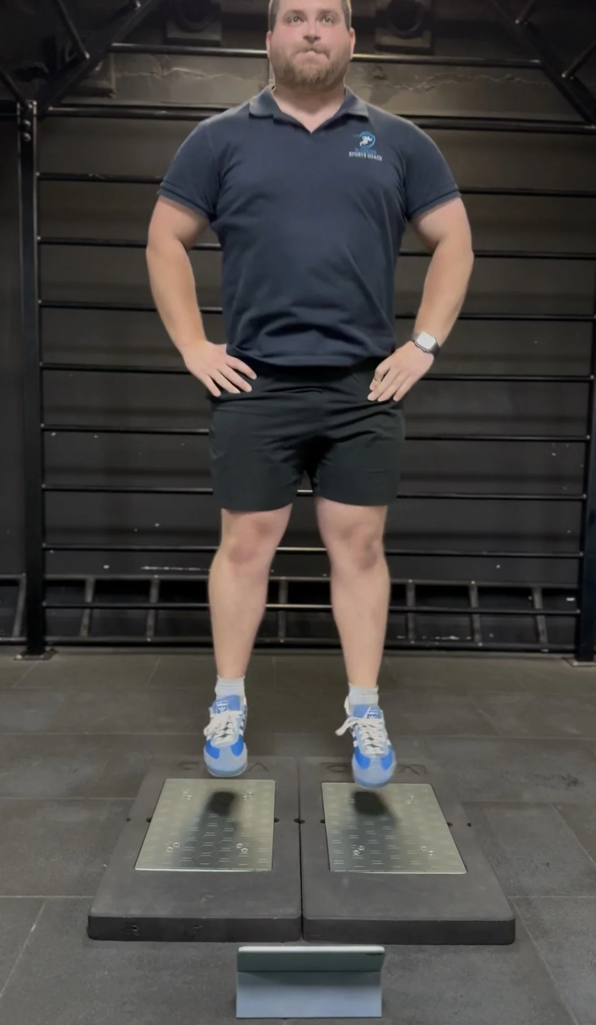 Man standing on force plates in a gym performing a jump for performance analysis, wearing a navy polo with a logo, black shorts, blue and white sneakers, and a smartwatch, with a black wall and gym equipment in the background.