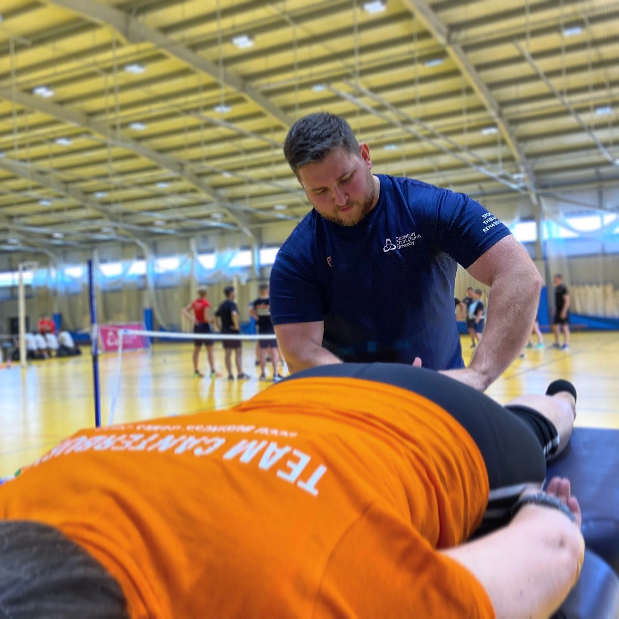A man preparing for a sport activity in an indoor sports arena, wearing a blue shirt with university logo, with an orange sports ball in front of him and other athletes in the background.