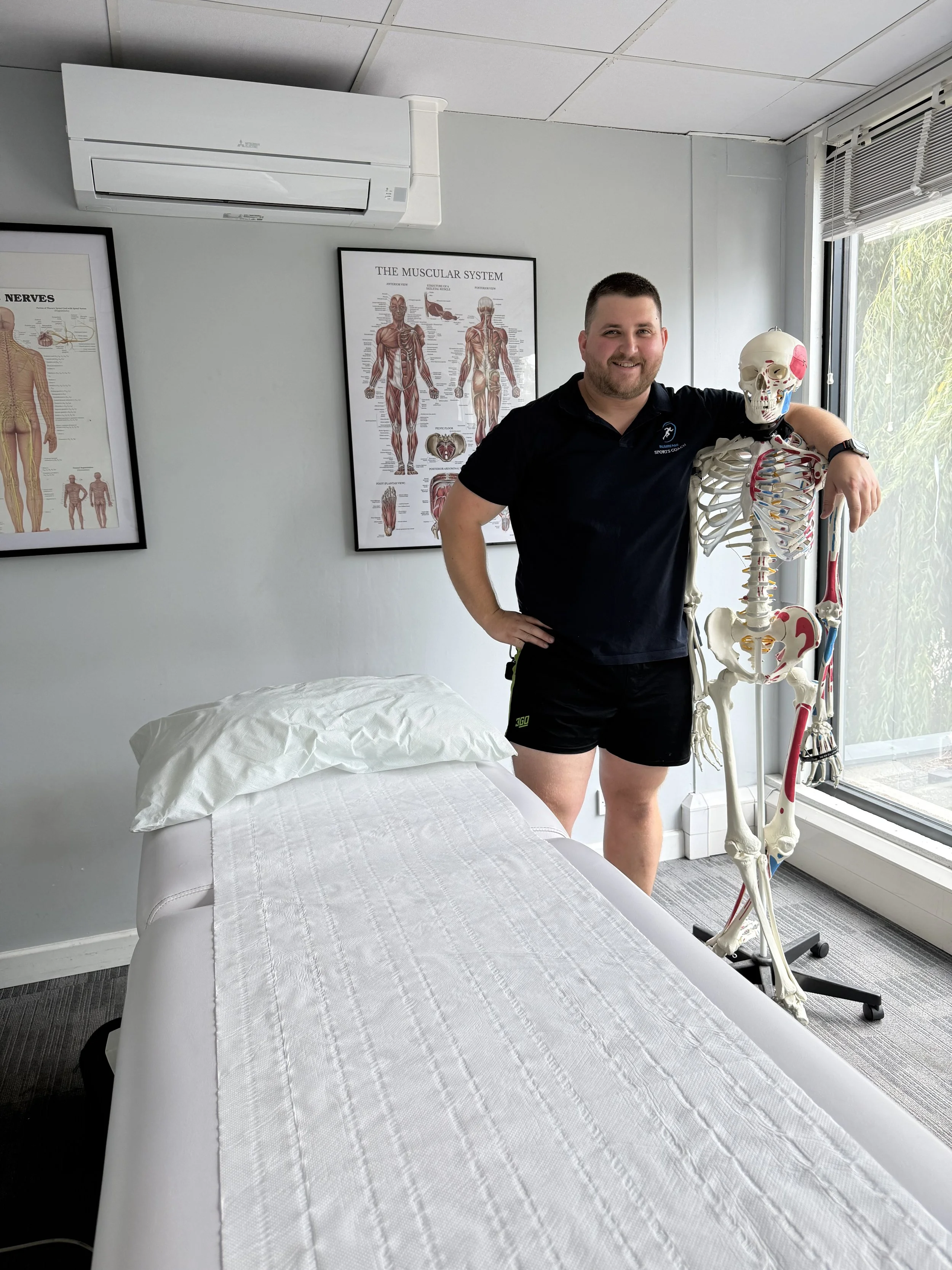 A man standing in a medical office with a muscular system poster and a life-sized skeleton model nearby, next to a bed covered with white sheets.