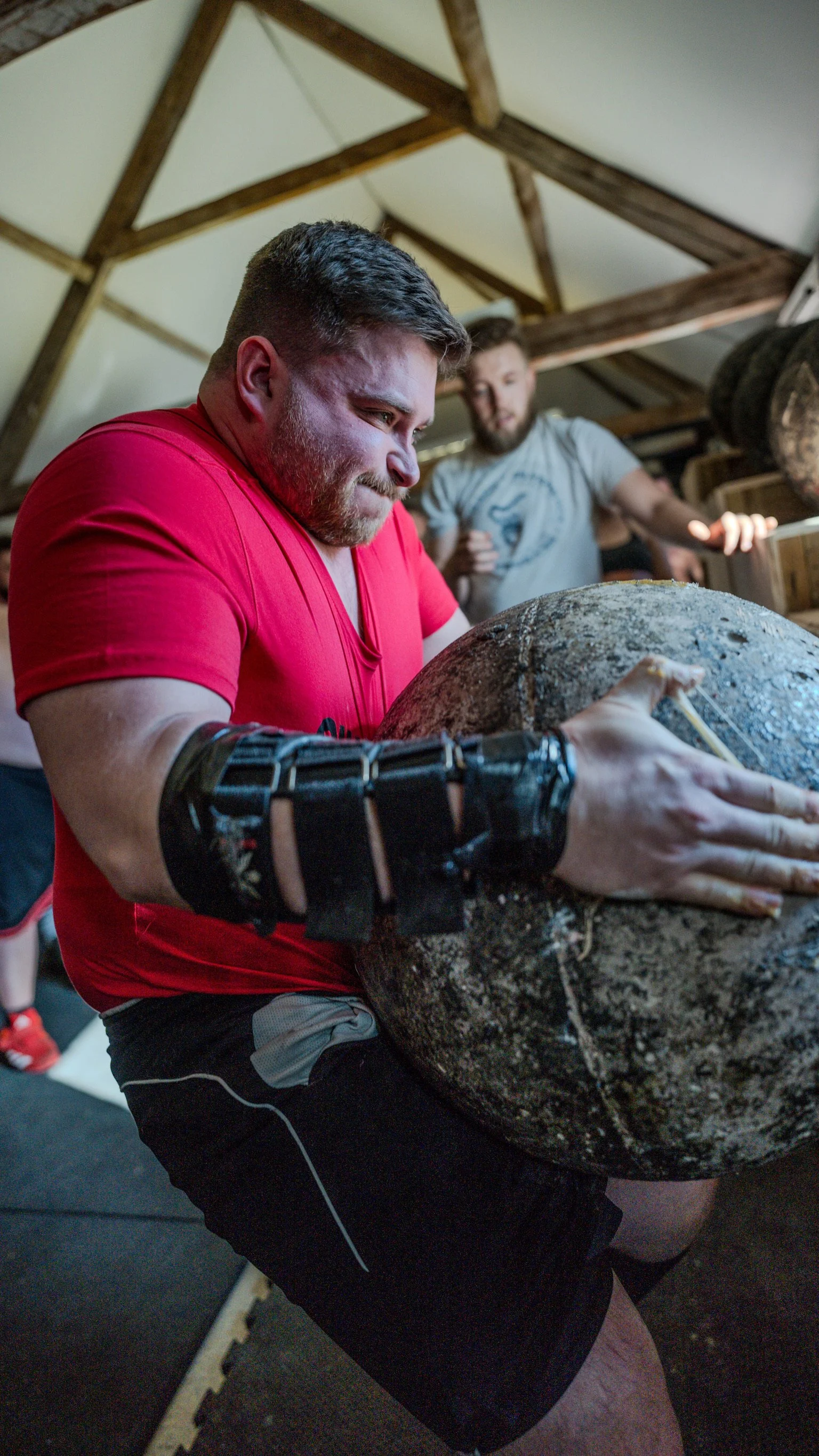 A man  lifting a large rock in a gym or training space, with another man in the background.
