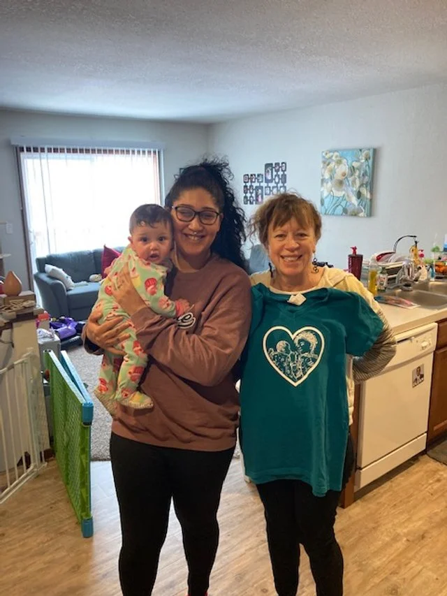 Two women smiling, one holding a baby, in a kitchen with a living room background. This is a family supported by In-Home Services through MomsBloom in Grand Rapids.
