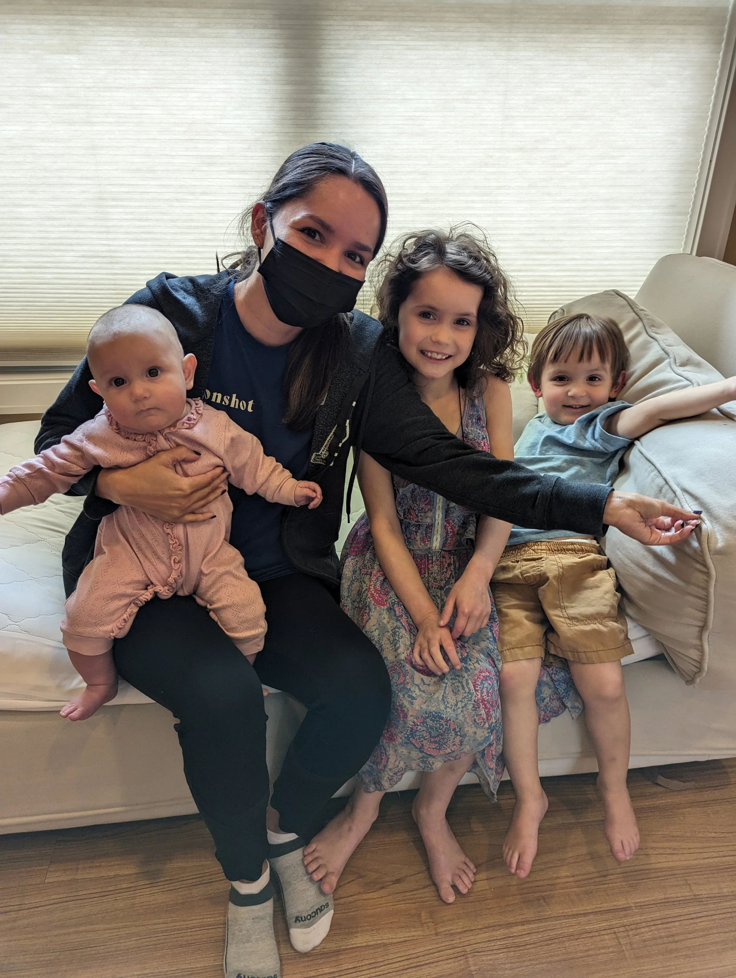 Four people sitting on a beige couch: an adult woman wearing a black face mask, a young girl with curly brown hair wearing a dress, a toddler boy with brown hair in a gray t-shirt and khaki shorts, and an infant girl in pink clothing. The woman is ho