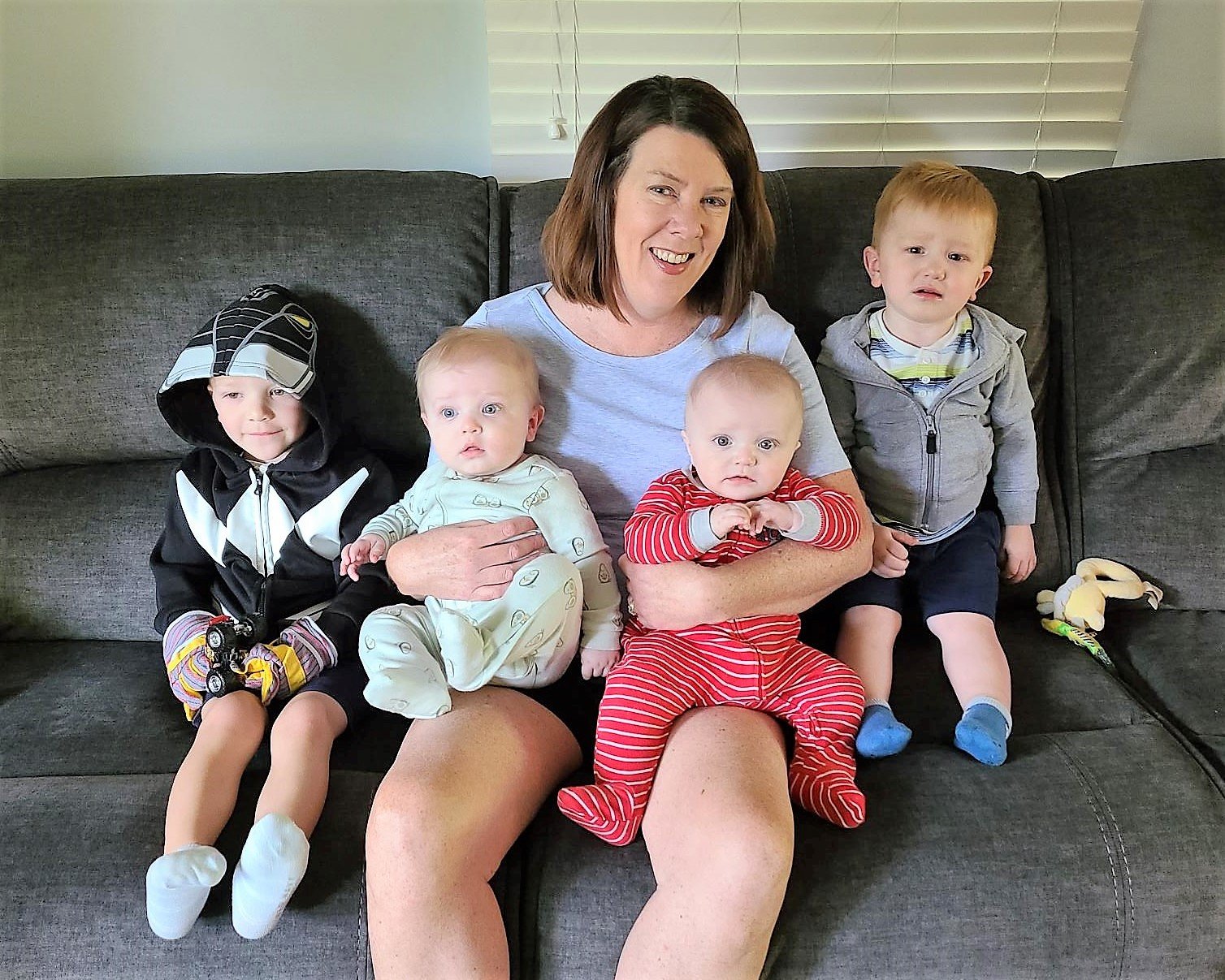 A woman sitting on a dark gray couch with four young children, some sitting on her lap and others beside her, in front of a window with blinds.