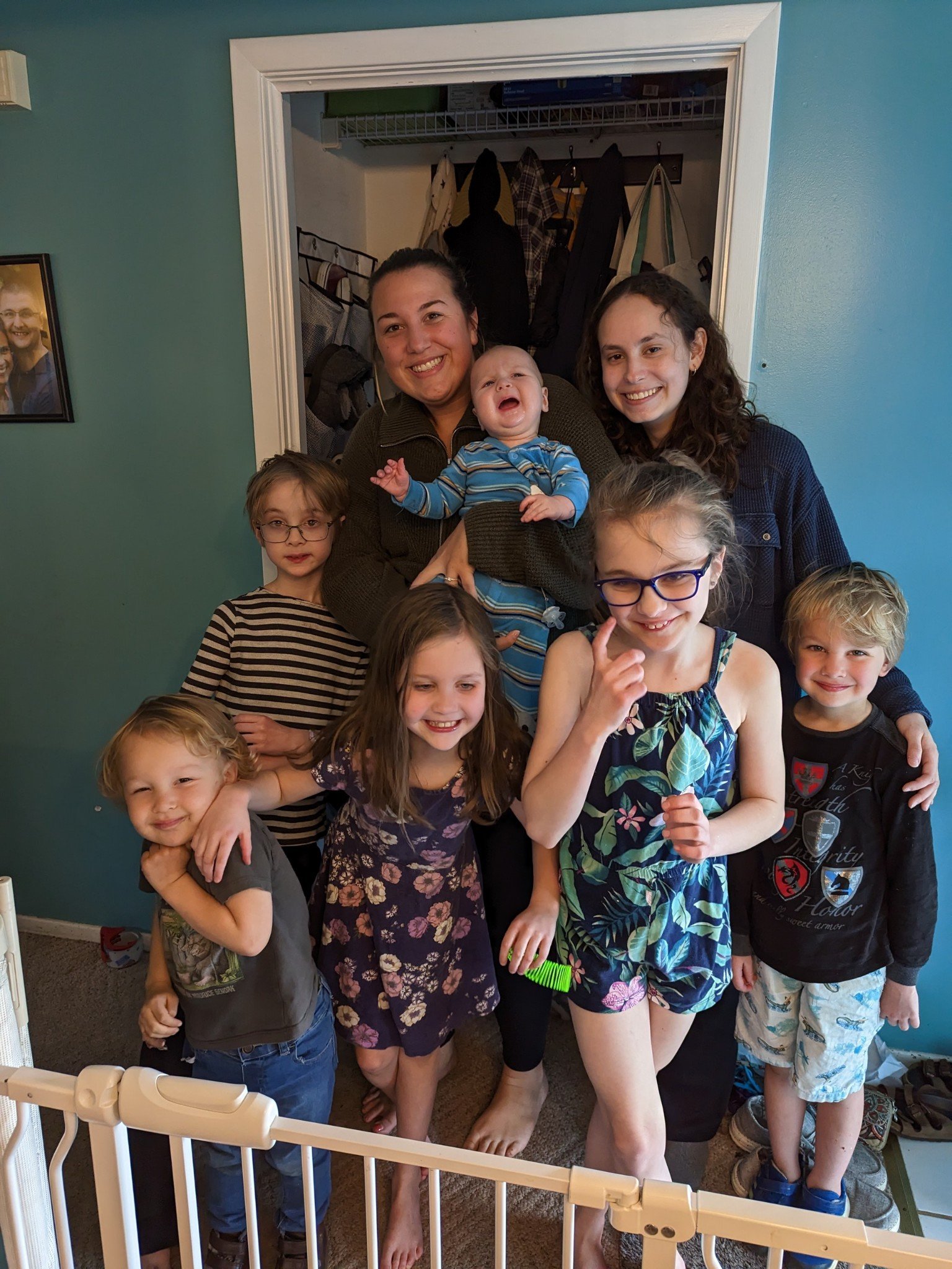 A group of five children and two adults smiling and posing together in a room, with a baby being held by one adult in front of a doorway.